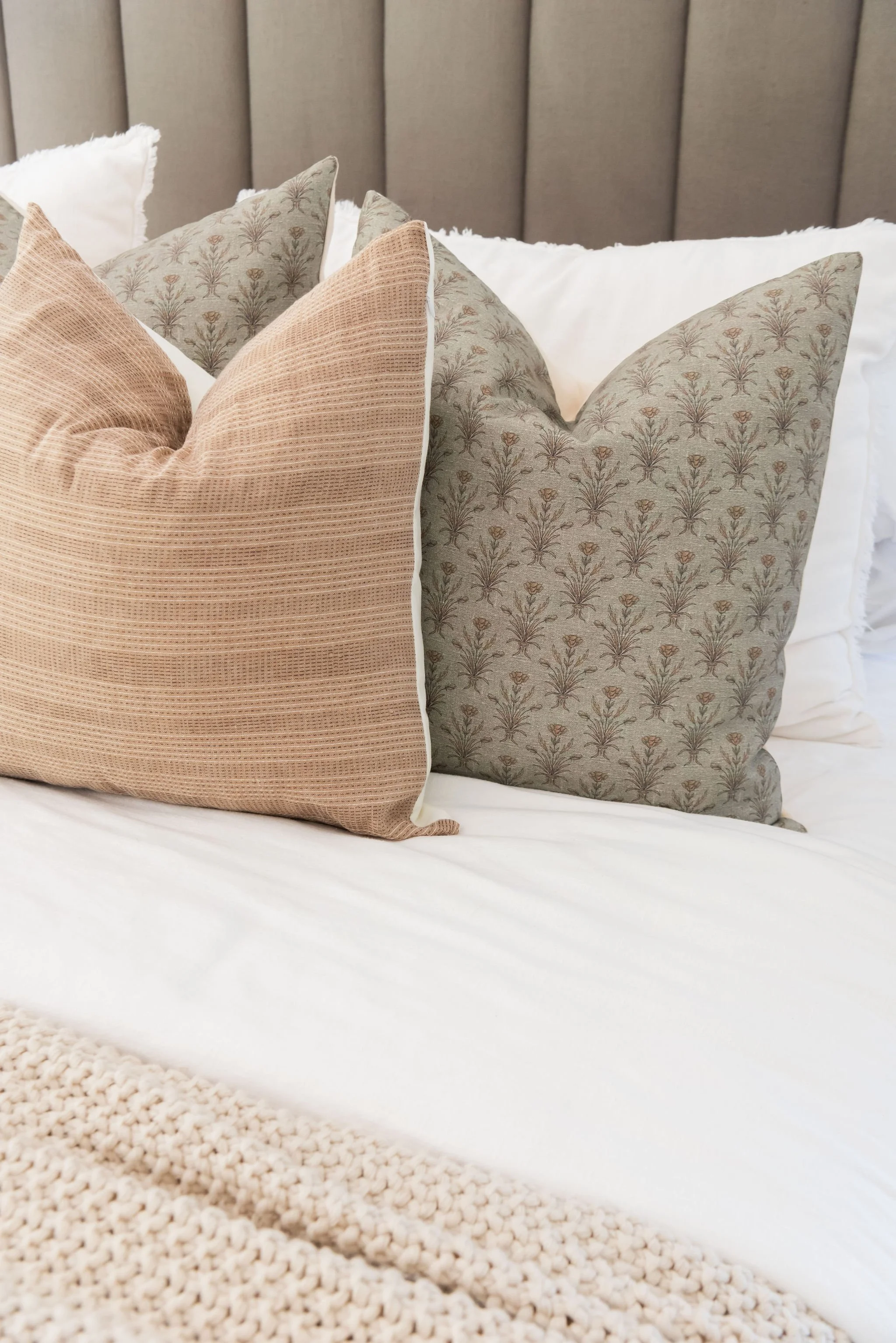 A close-up of a bed with multiple decorative pillows, including a beige textured pillow and two gray pillows with a floral pattern, on a white bedspread with a knitted blanket at the foot of the bed.