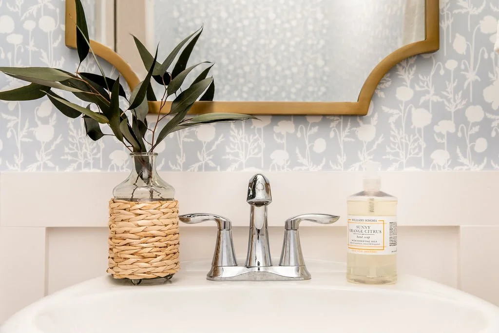A bathroom sink with a chrome faucet, a clear bottle of hand soap labeled "Sunny Orange Citrus," and a glass vase with green leafy branches in a woven holder, all reflected in a mirror with a gold-colored frame.