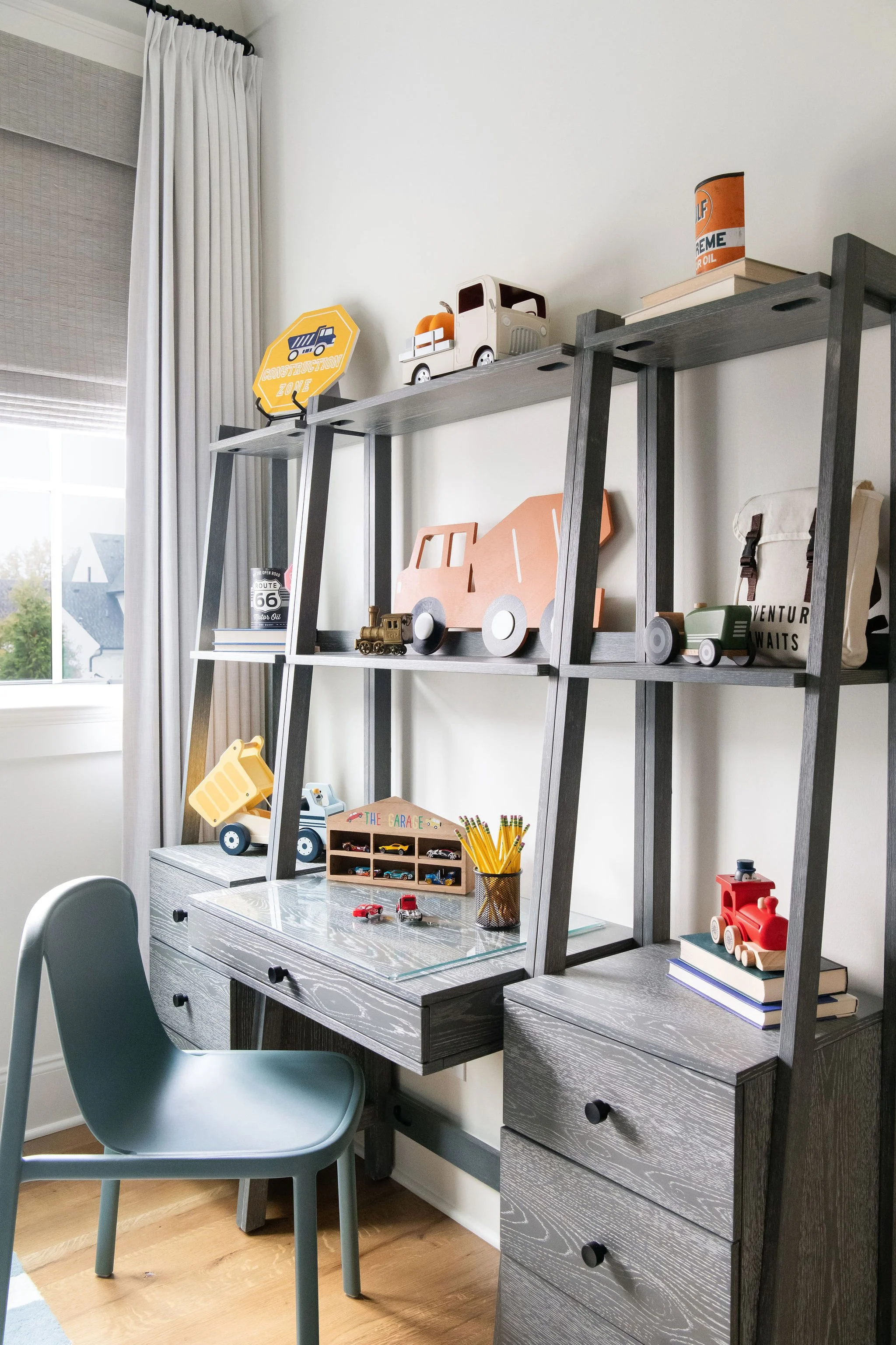 A desk with a ladder-style bookshelf behind it, decorated with toy vehicles, books, a pencil holder, and a small car model; a blue chair is in front of the desk, and a window with curtains is to the left.