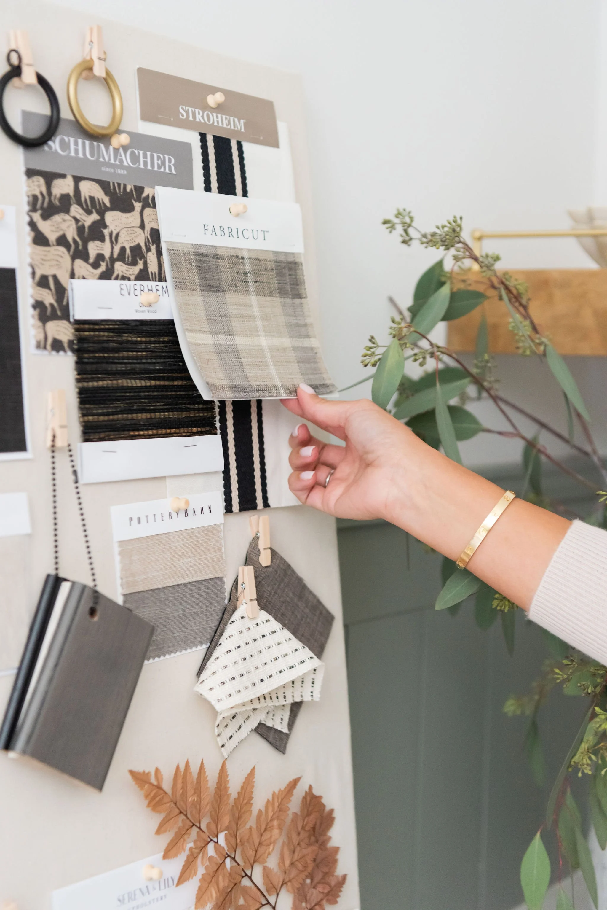 Person holding a fabric swatch in front of a display board with various fabric and material samples, and some decorative leaves on a table.