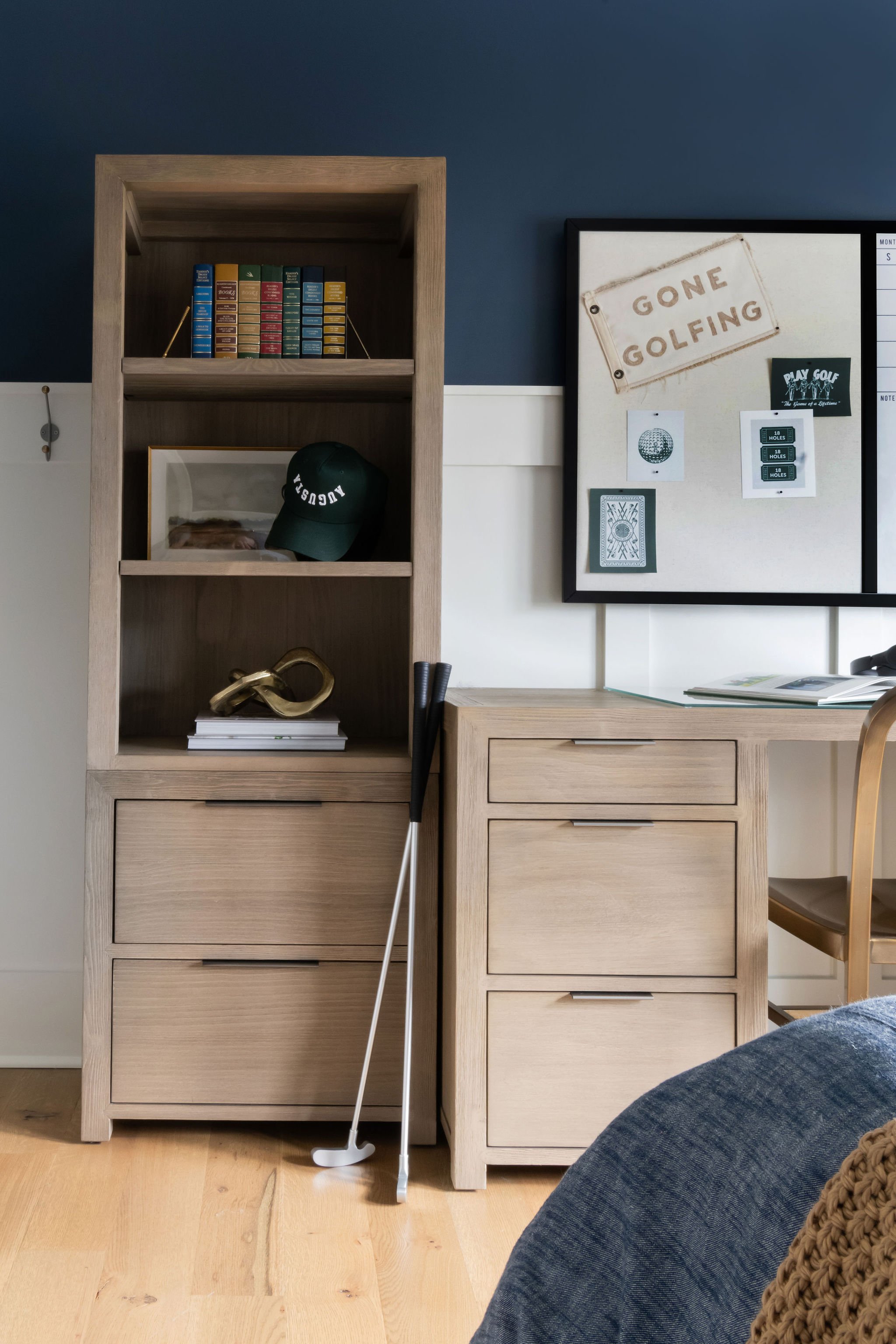 A modern interior with wooden furniture, a bookshelf with colorful books and a black cap, golf clubs, and decor. A sign on the wall says 'GONE GOLFING' with various golf-related papers and a small chair nearby.