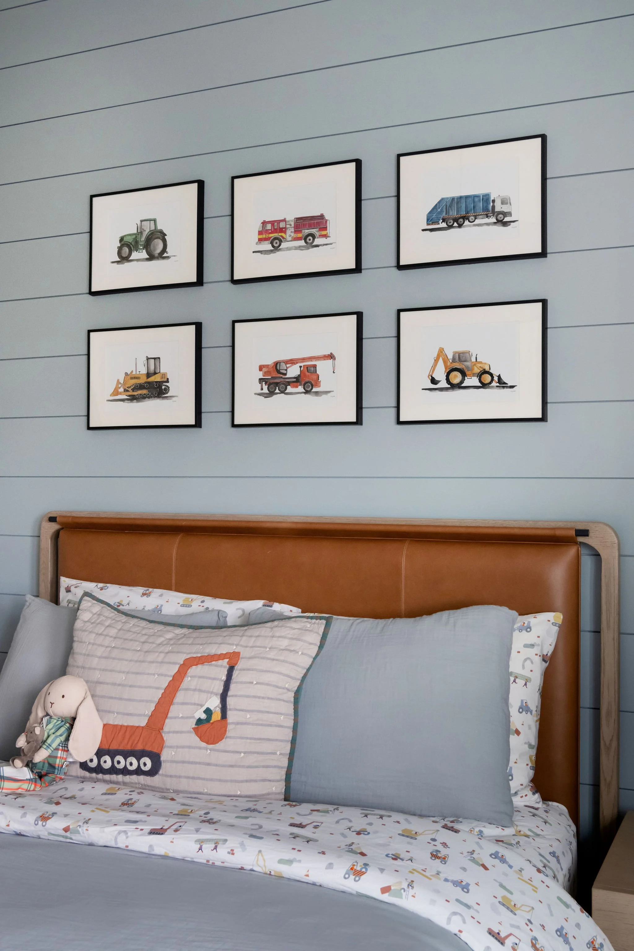 Bedroom with framed pictures of construction vehicles on the wall above the bed, which has a leather headboard and bedding with construction vehicle patterns.