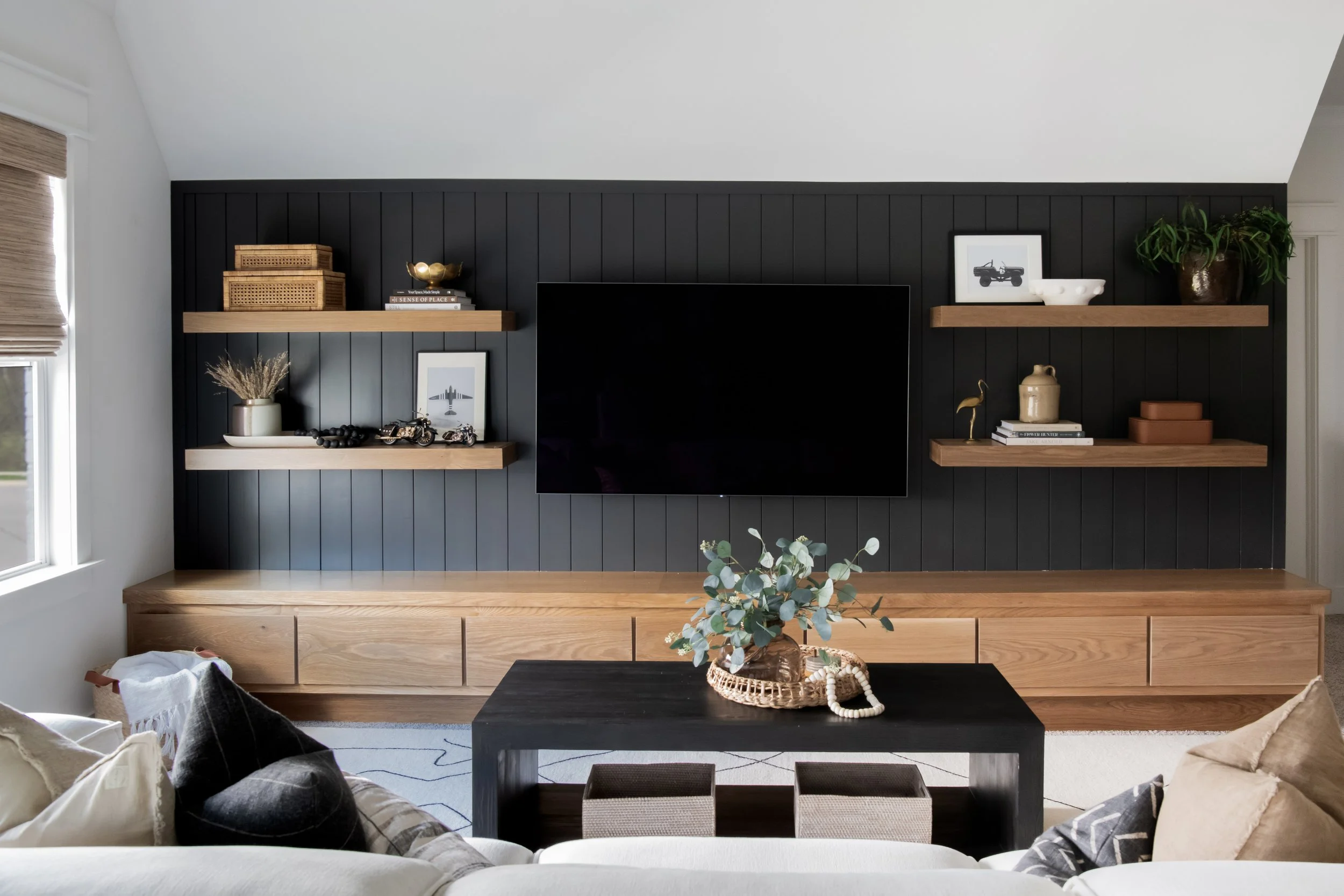 Living room with black accent wall, mounted flat-screen TV, wooden shelves with decorative items, a white sofa with pillows, a black coffee table with a plant and jewelry, and a window with a woven shade.