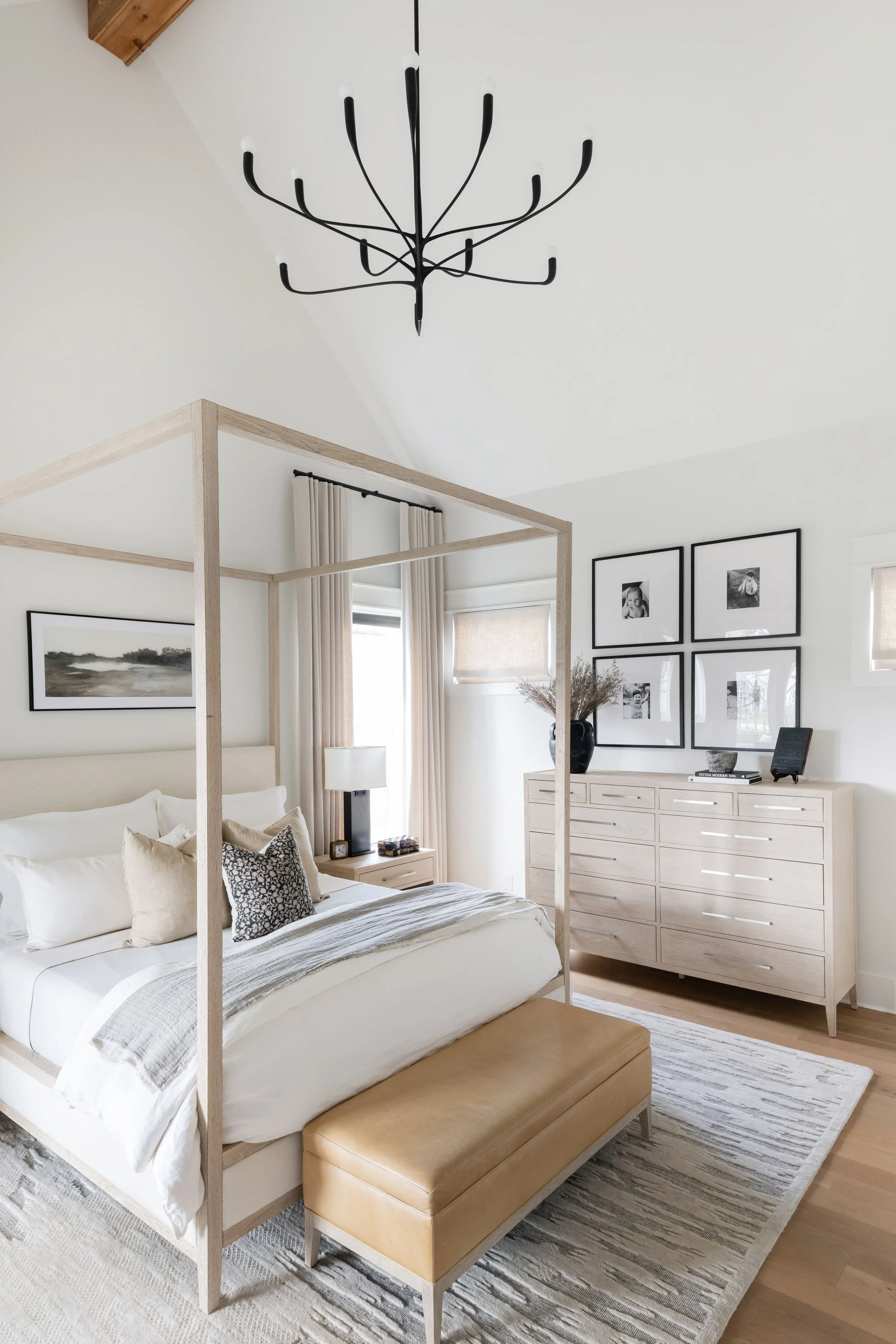 A contemporary bedroom with a four-poster bed, beige upholstered bench, light wood dresser, framed black-and-white photos on the wall, and a black chandelier hanging from the ceiling.