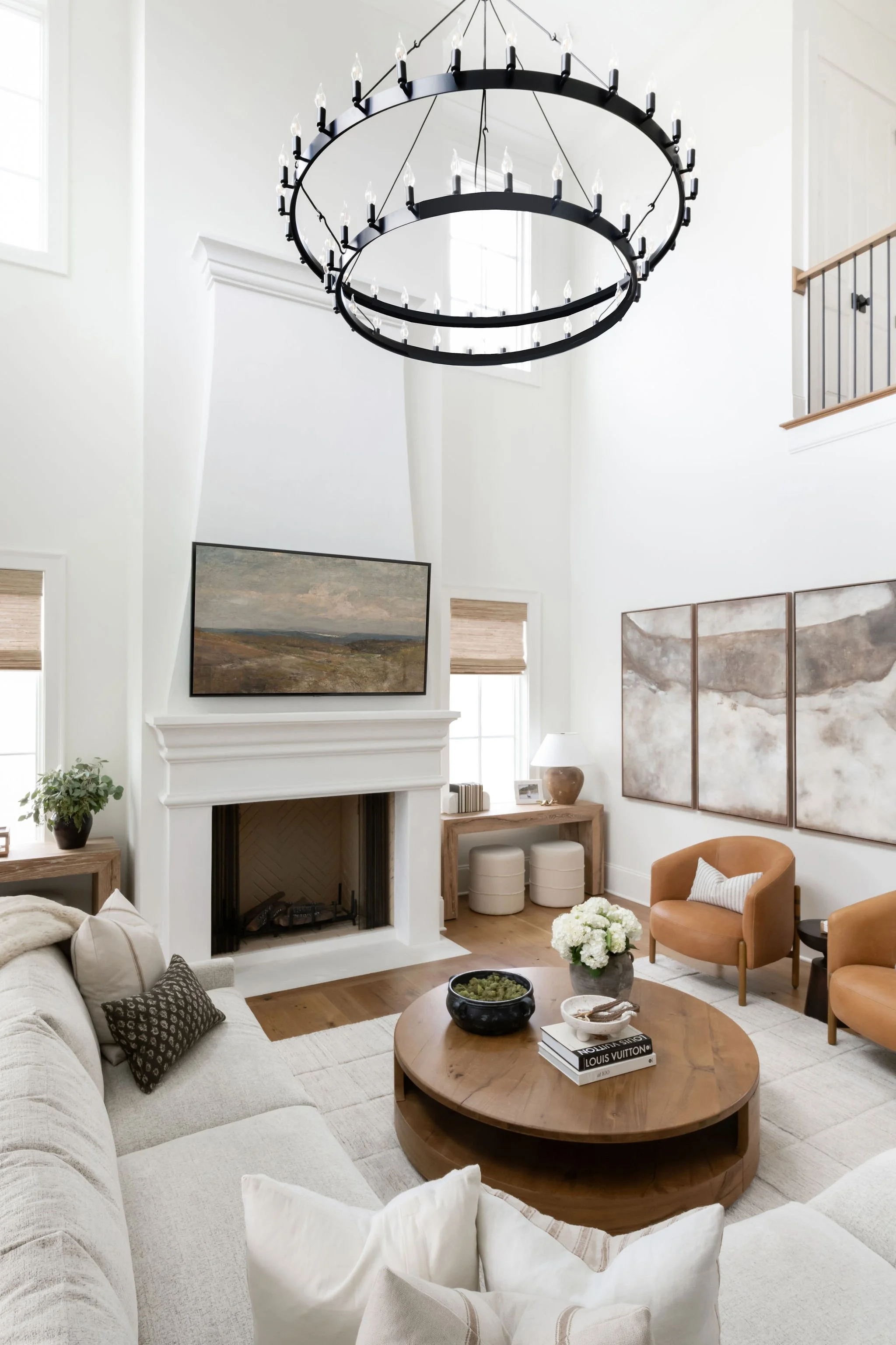 Bright living room with white walls, a large round black chandelier, a fireplace with a TV above, and beige and tan seating, including a sectional sofa and two armchairs. Decor includes a wooden coffee table, books, paintings, and a potted plant.