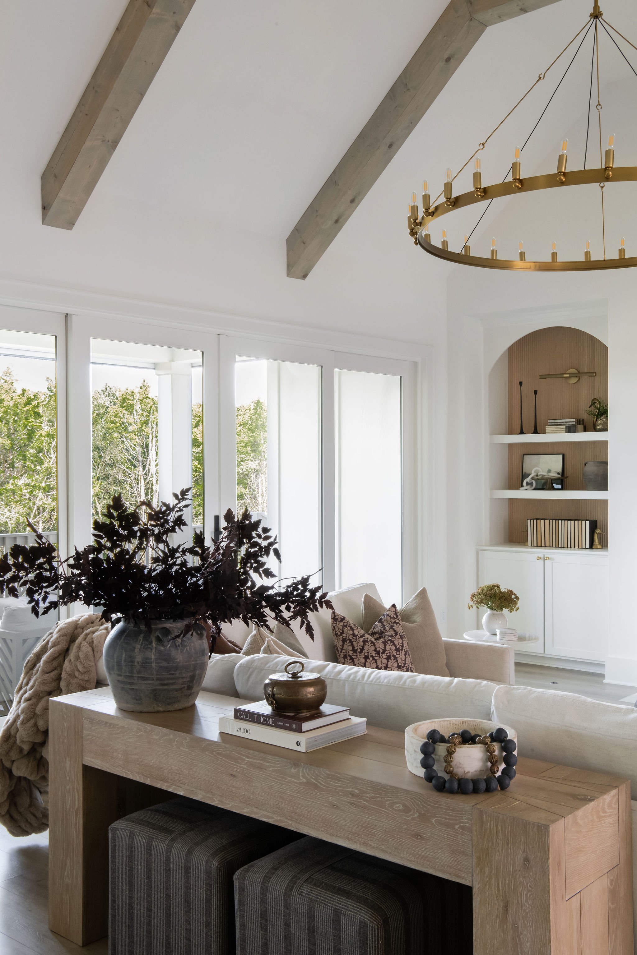 A bright living room with large windows, a white sofa with multiple pillows, a wooden table with decorative items, a shelf with books and decor, and a chandelier hanging from the ceiling with wood beams.