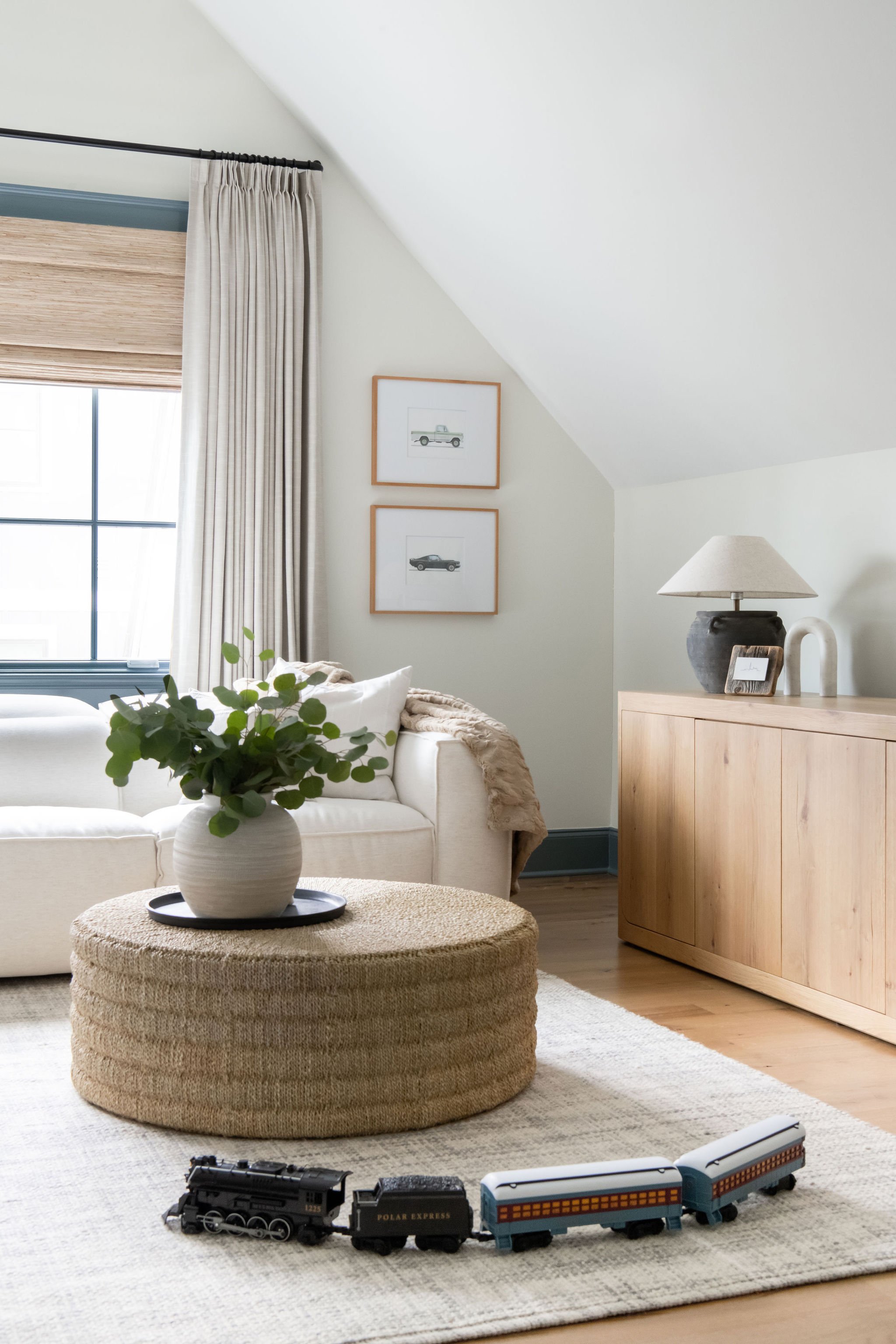 Living room with cream sofa, round woven ottoman with a vase of greenery, wooden sideboard with lamp and decorative items, framed car prints on wall, window with curtains, and toy train on rug.