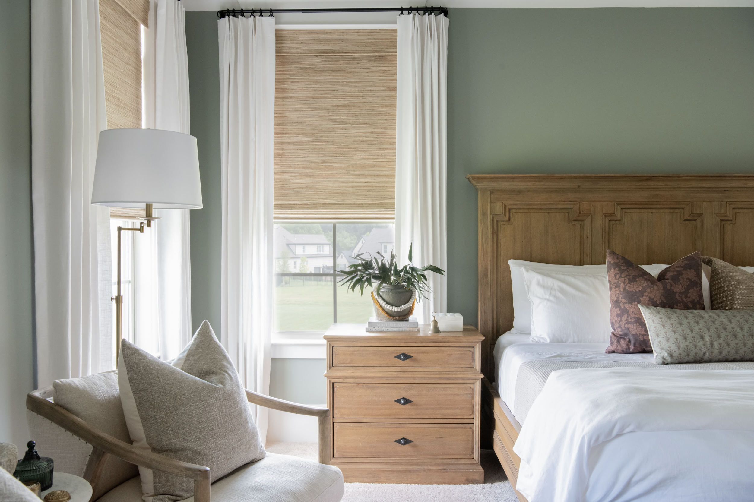 A bedroom with a wooden bed, white bedding, and decorative pillows. A wooden nightstand with a potted plant and books, and a cozy armchair with pillows. Large windows with white curtains and a view of houses and greenery outside.