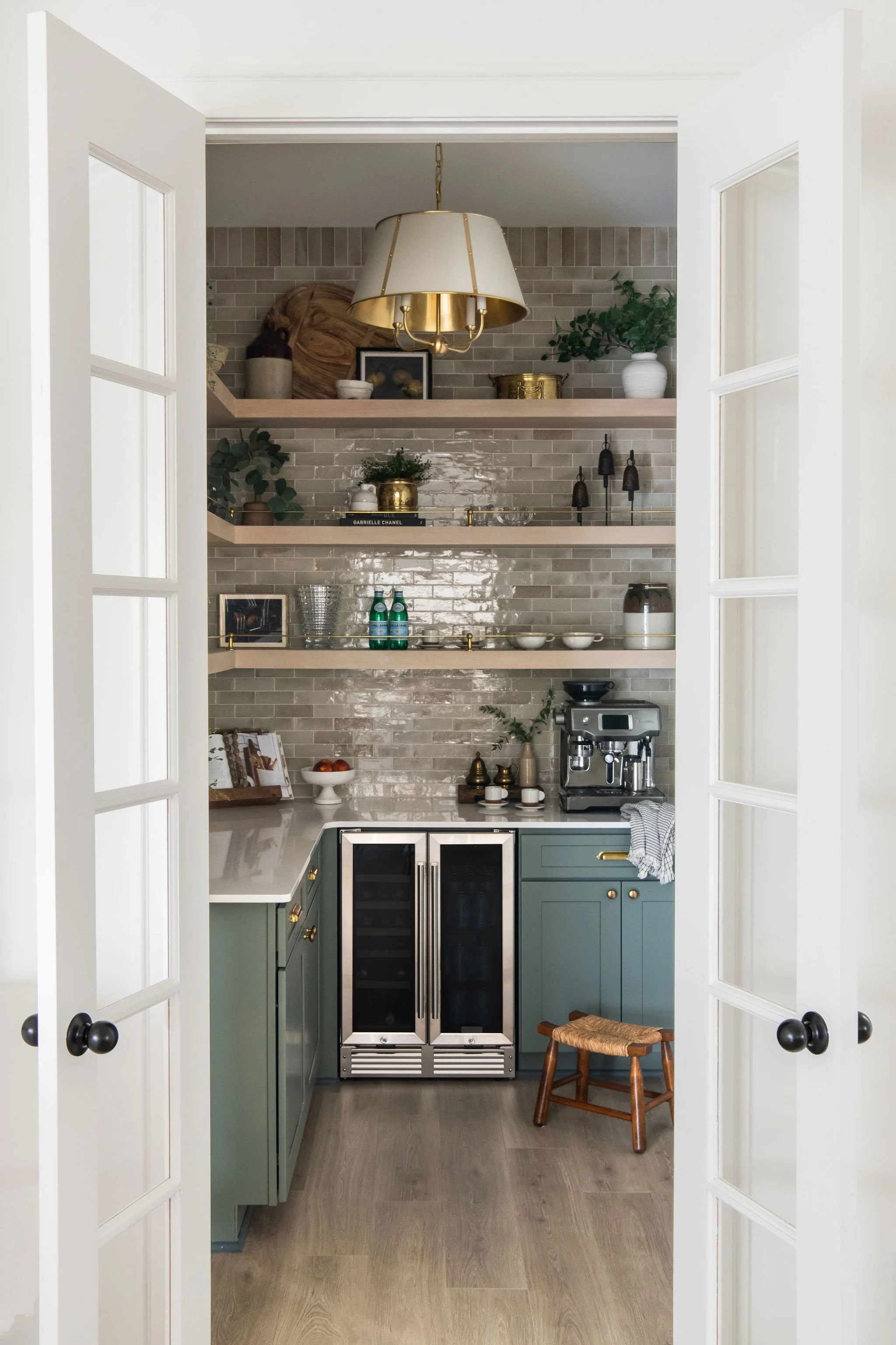 View of a modern kitchen seen through white double doors, with blue-green cabinets, open shelves with decor, and a tiled backsplash.