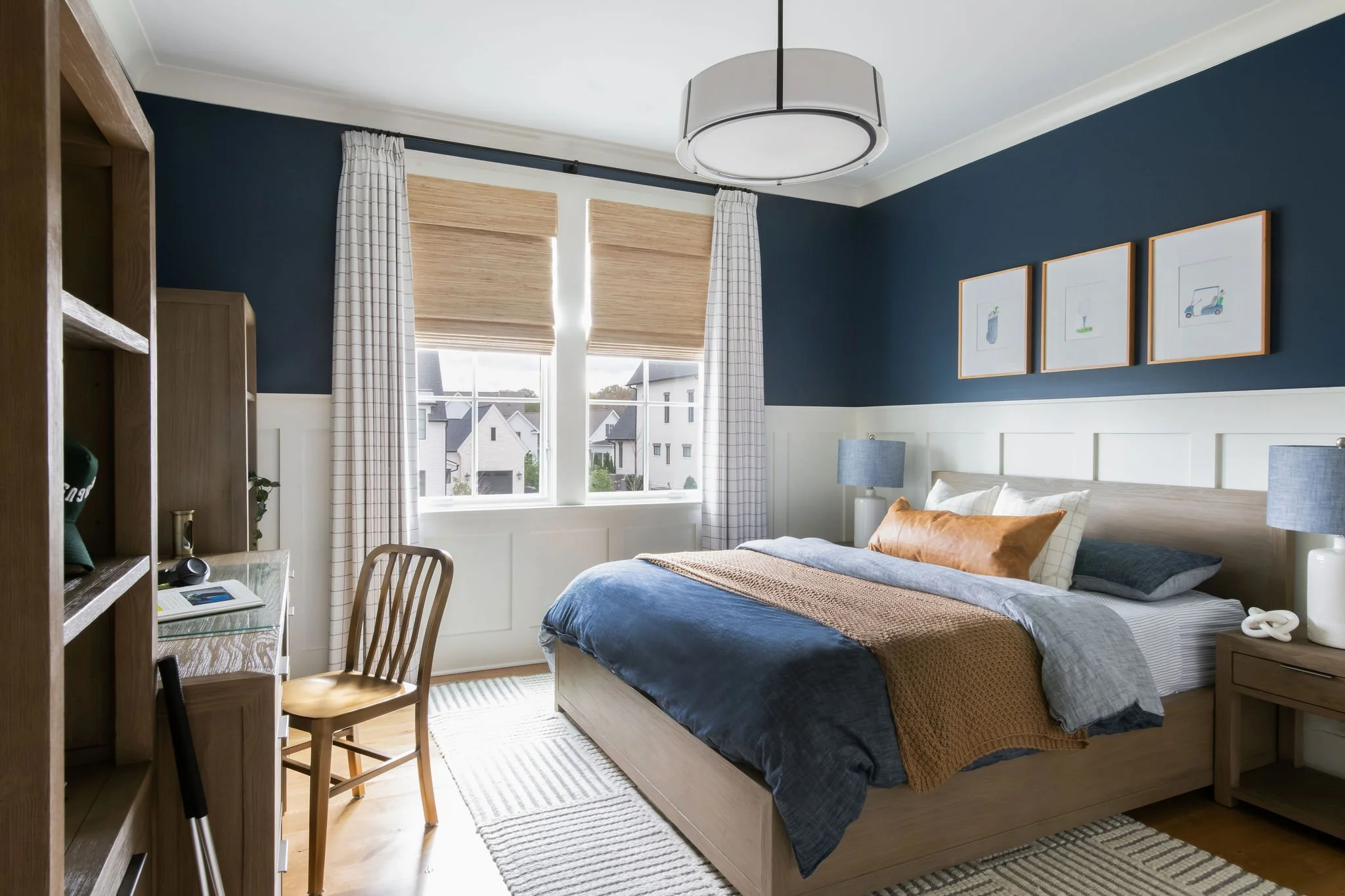 A modern bedroom with a large bed, navy and neutral bedding, and artwork of simple plants and objects on a navy accent wall. There are two bedside tables with lamps, a bookshelf, a desk with a chair, a window with blinds and curtains, and light wood 