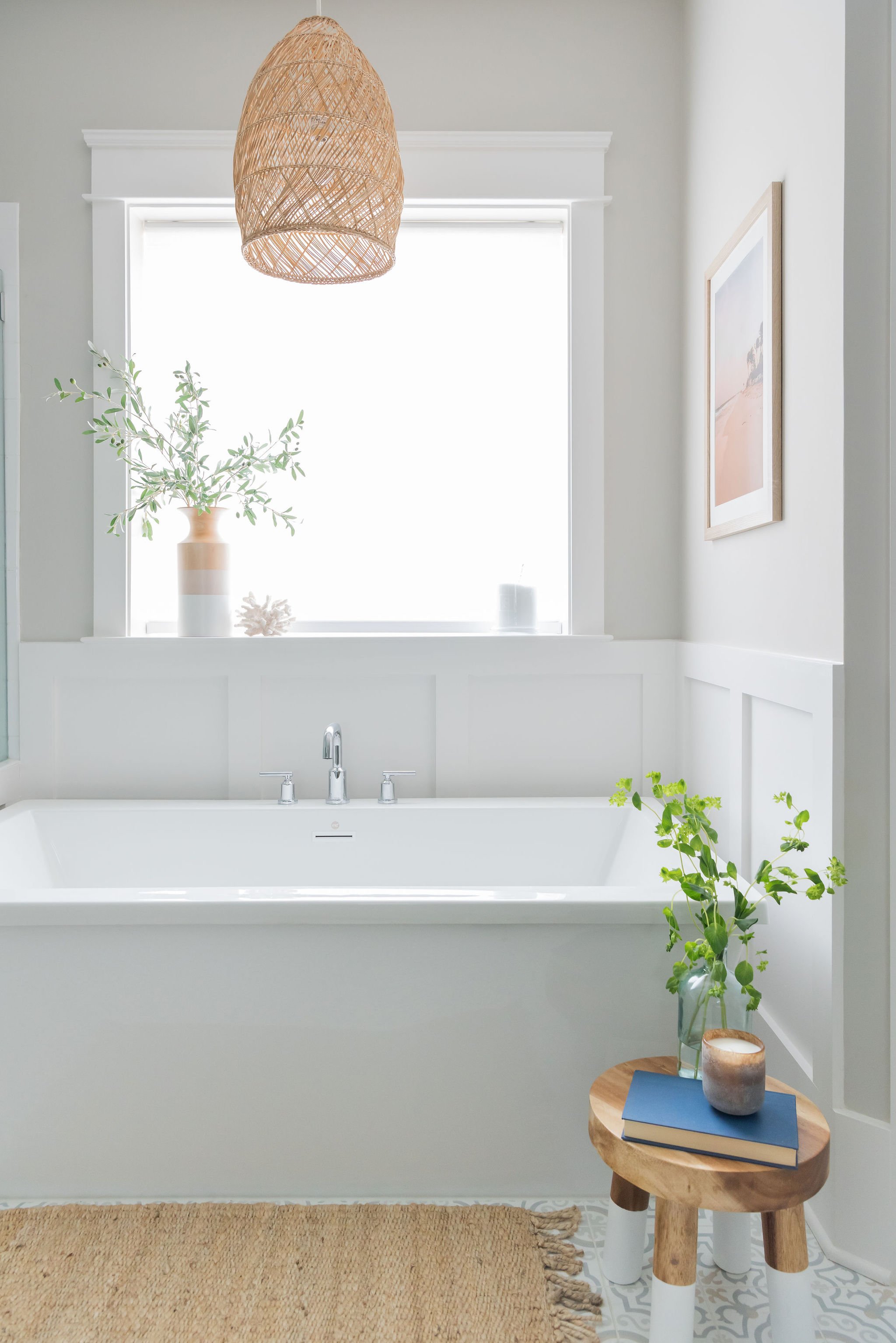 Bright bathroom with a white bathtub, natural light from a large window, plants, framed artwork, woven pendant light, and a small wooden stool with books and a candle.