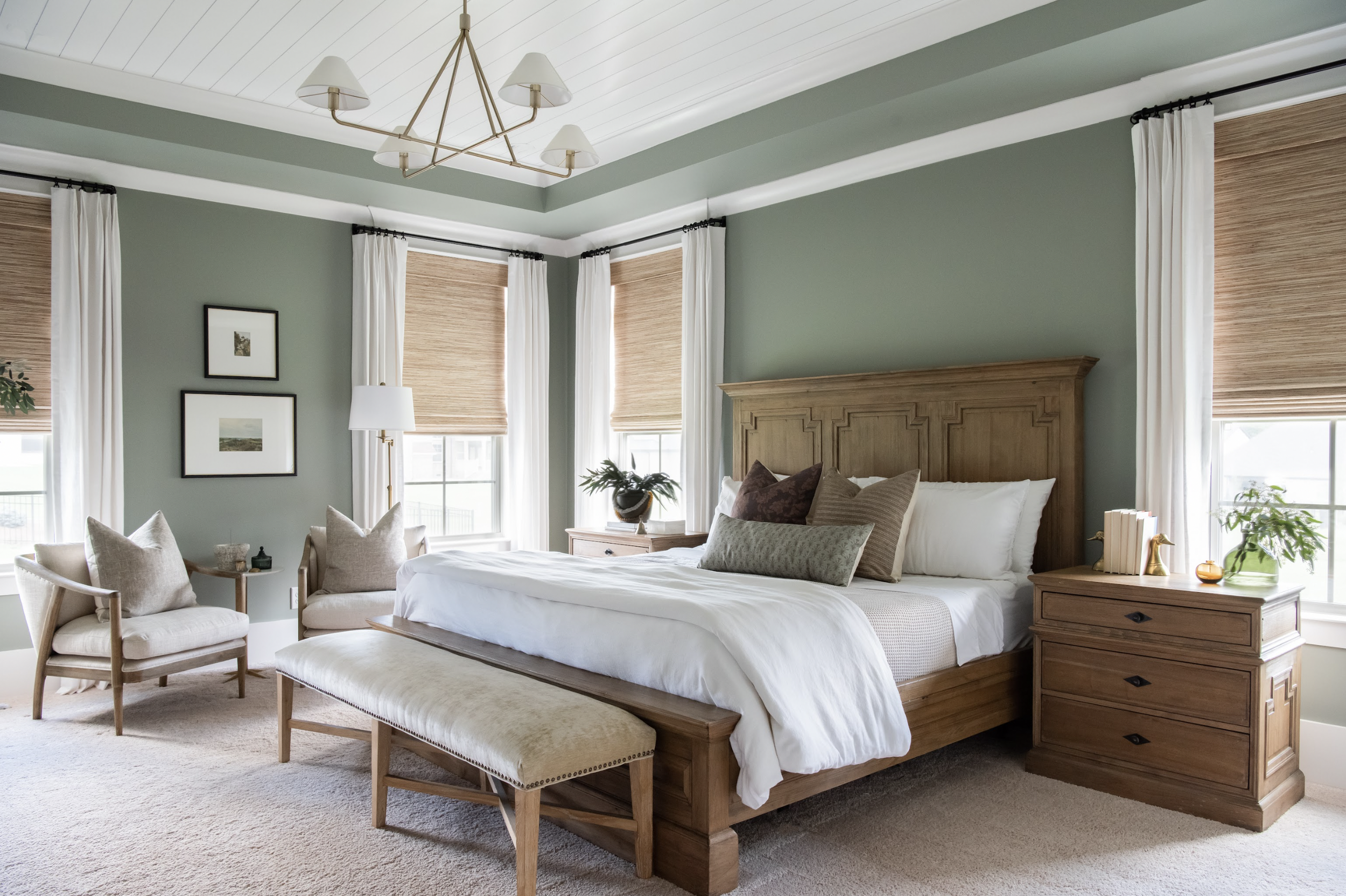 A bedroom with a large wooden bed, multiple pillows, a bench at the foot, two nightstands with plants and books, three windows with white curtains and bamboo blinds, a chandelier, two armchairs with pillows, a side table, and framed artwork on the wa