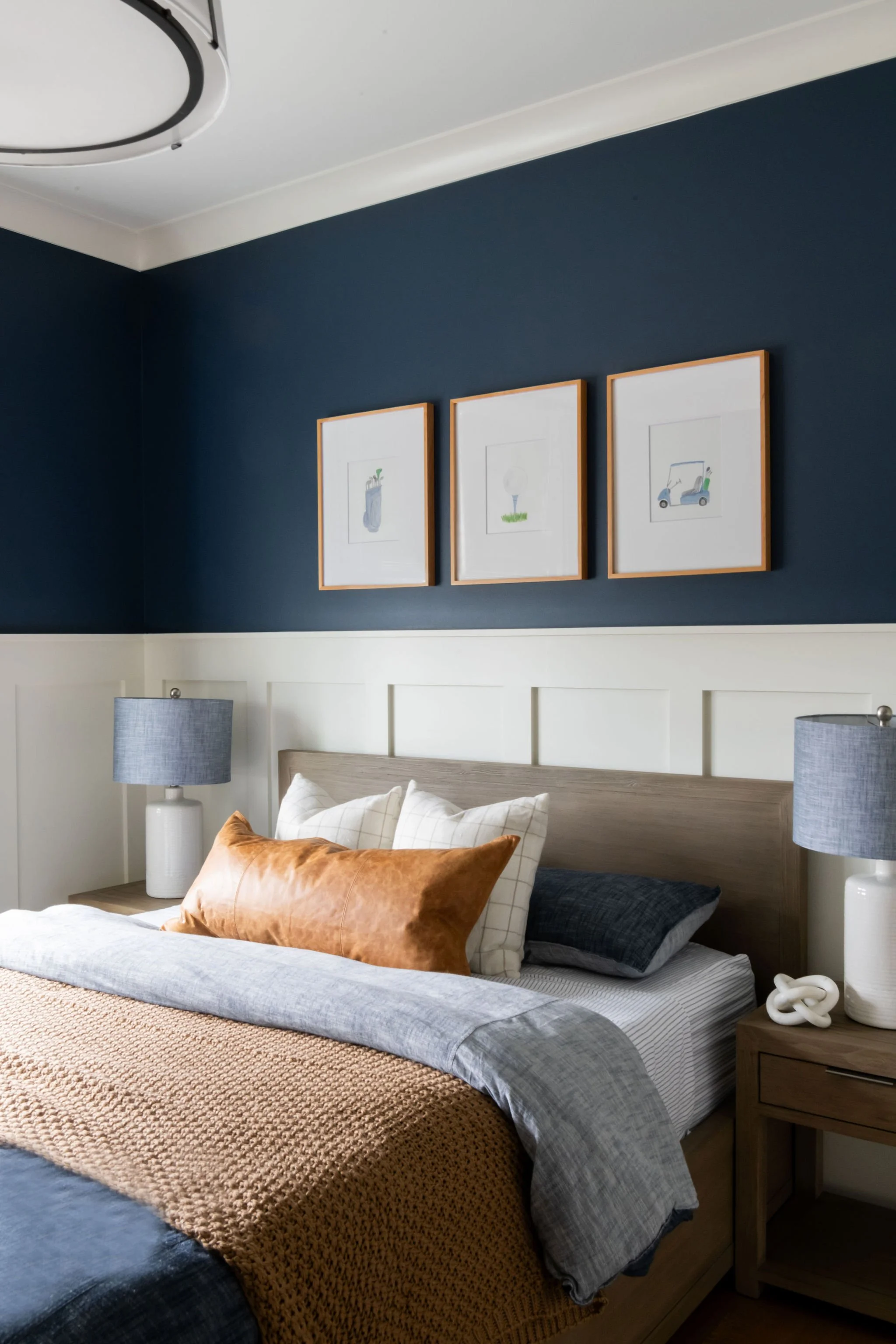 A neatly made bed with multiple pillows, including white, dark gray, and a brown velvet pillow, in a bedroom with navy blue and white walls, framed artwork above the bed, and two matching lamps on bedside tables.