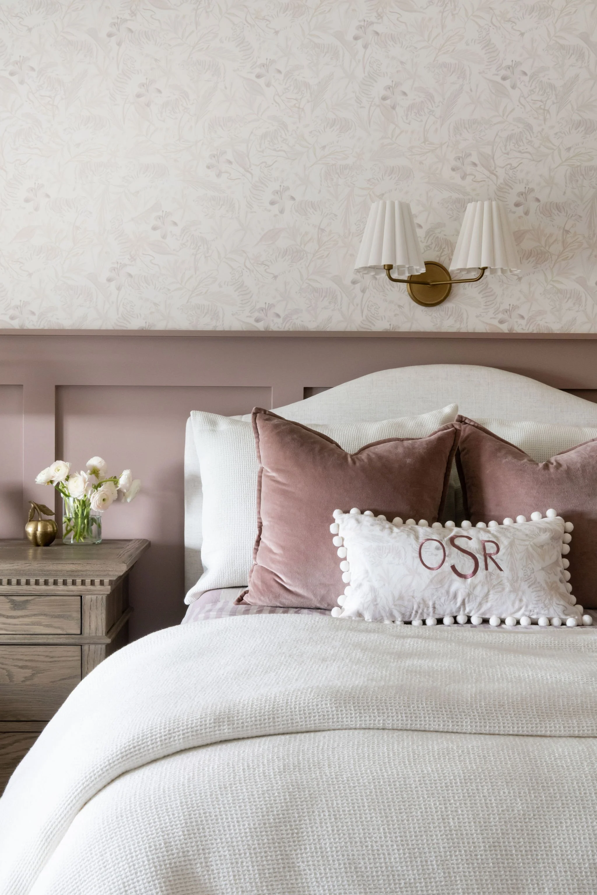 A bedroom with a white upholstered headboard, beige wall lamps, and decorative pillows on a bed. There are pink and white pillows, one embroidered with 'OSR', and a wooden nightstand with a small vase of white flowers.