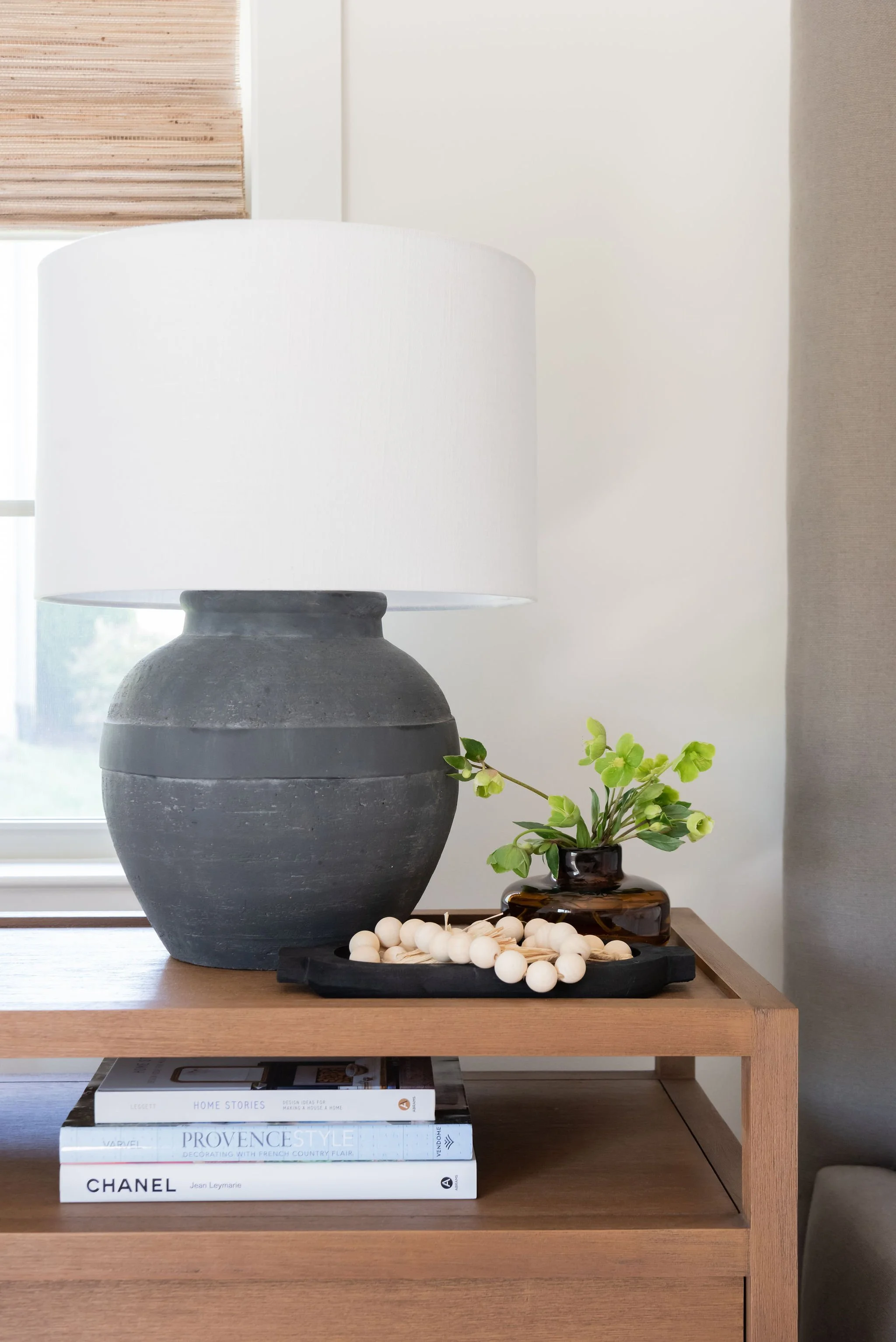 Decorative table lamp with a white shade on a wooden table, companioned by a small green plant, black tray with white beads, and a stack of books underneath the table.