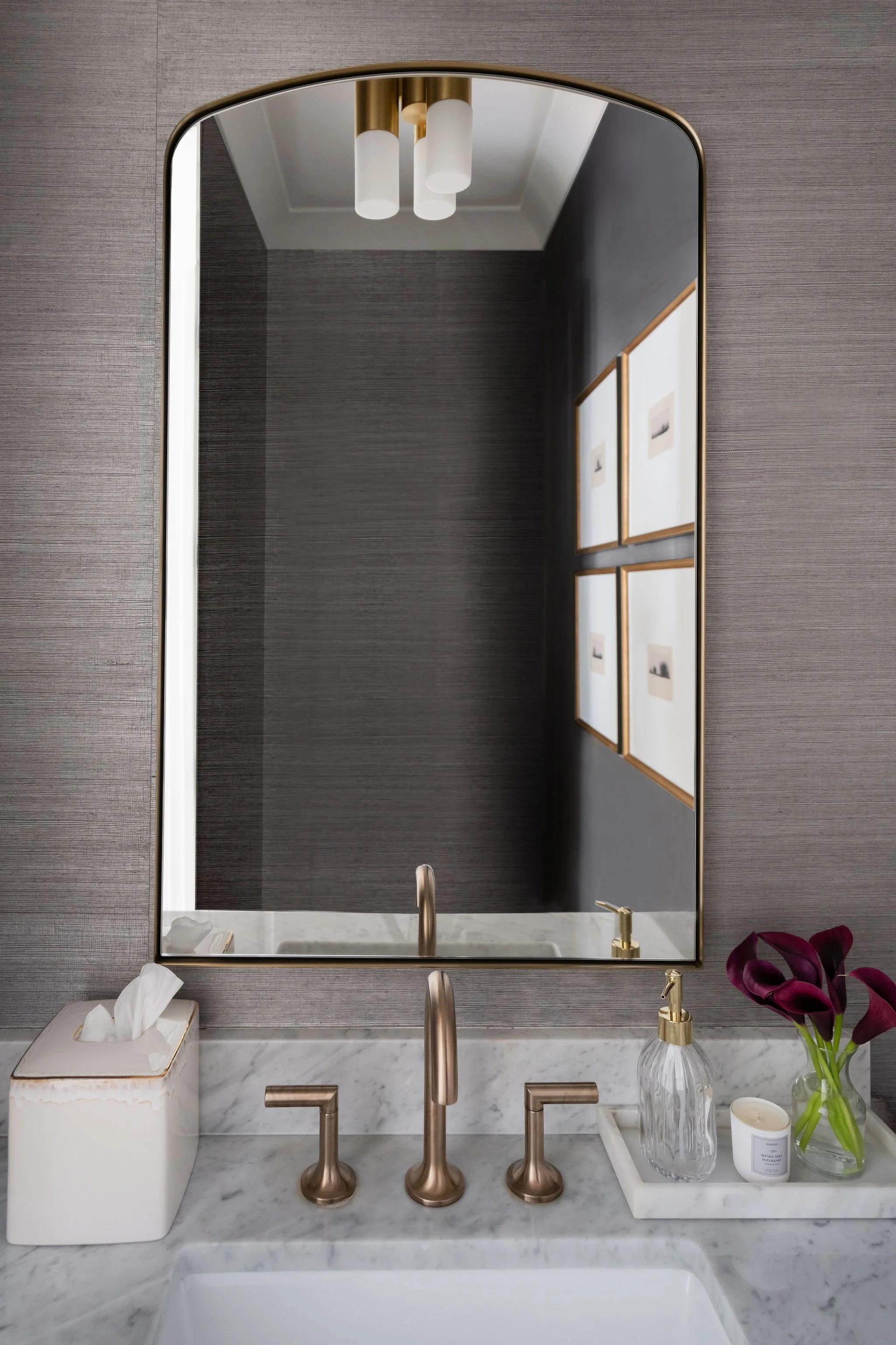 Bathroom vanity with a marble countertop, a large rectangular mirror, and wall-mounted faucet with two handles. Decorative items include a tissue box, a glass soap dispenser, a candle, and a vase with dark purple calla lilies. Four framed pictures ar