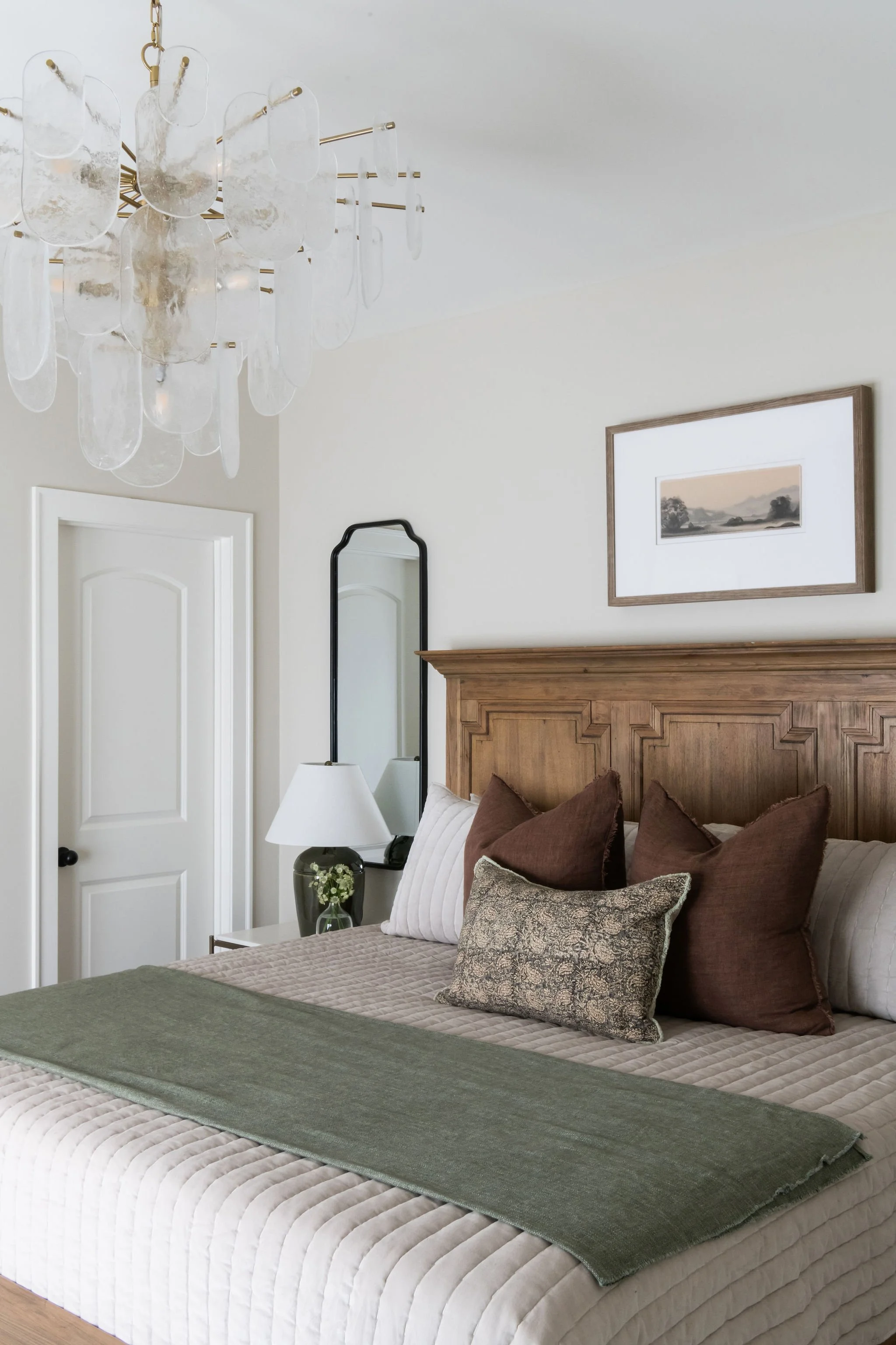 A bedroom with a wooden headboard, beige bedding, multiple pillows, a bedside table with a lamp, a mirror, a framed landscape picture, and a chandelier with glass elements.