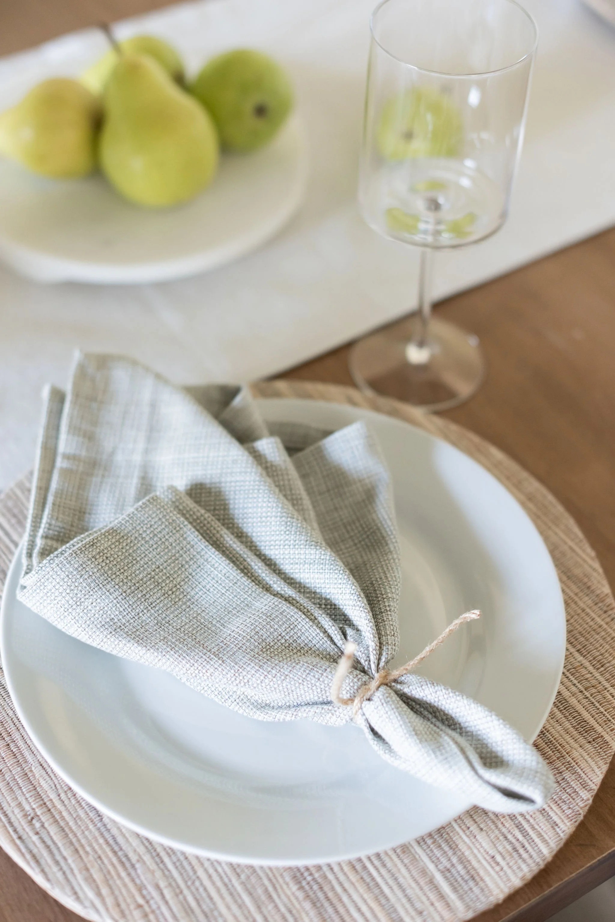 A table setting with a white plate, a folded cloth napkin tied with string, a clear empty wine glass, and a plate of green pears on a white tablecloth.