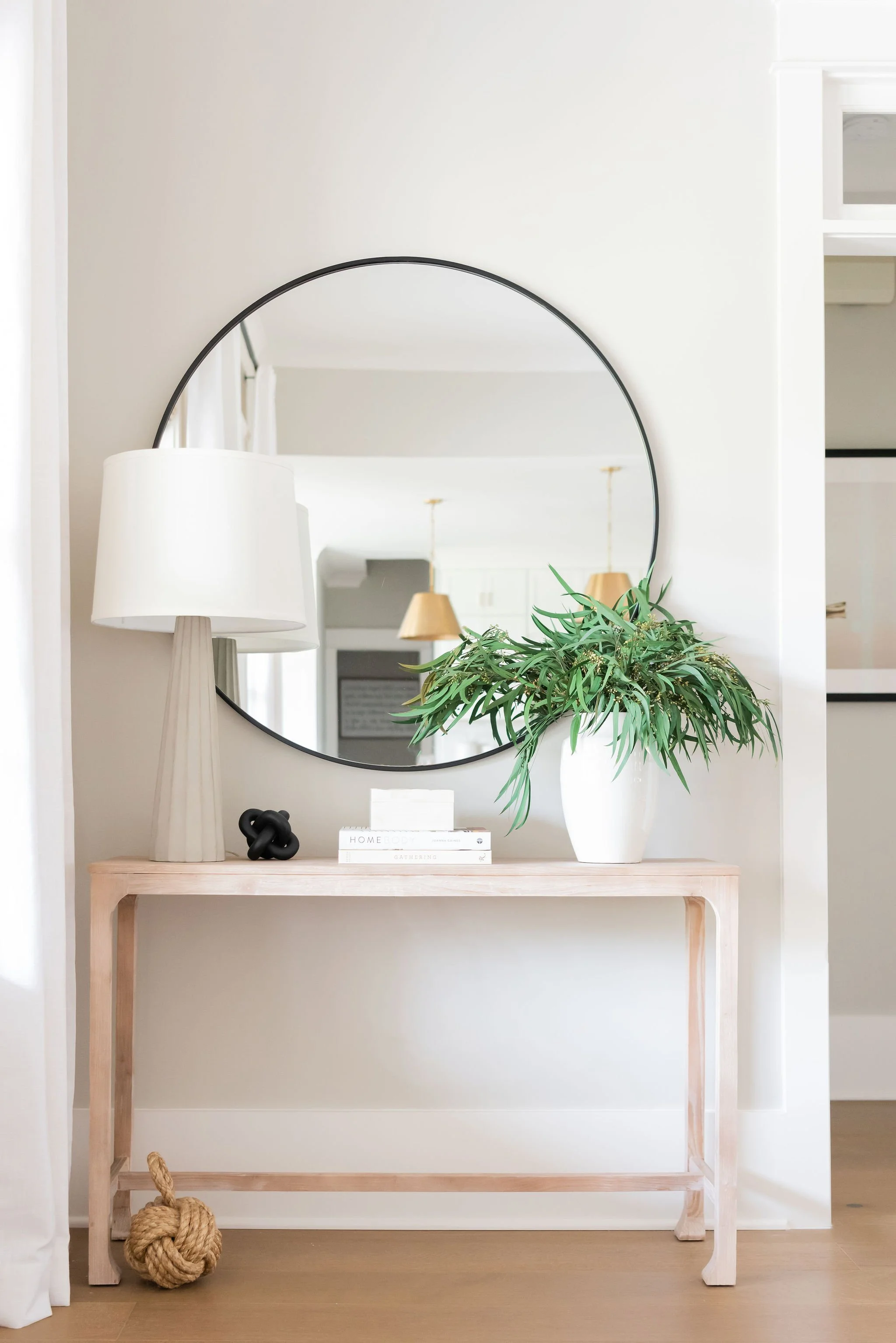 A minimalist entryway with a light wood table, a round mirror, a white lamp, a white vase with greenery, a small black sculpture, and books, with a hanging light fixture reflected in the mirror.
