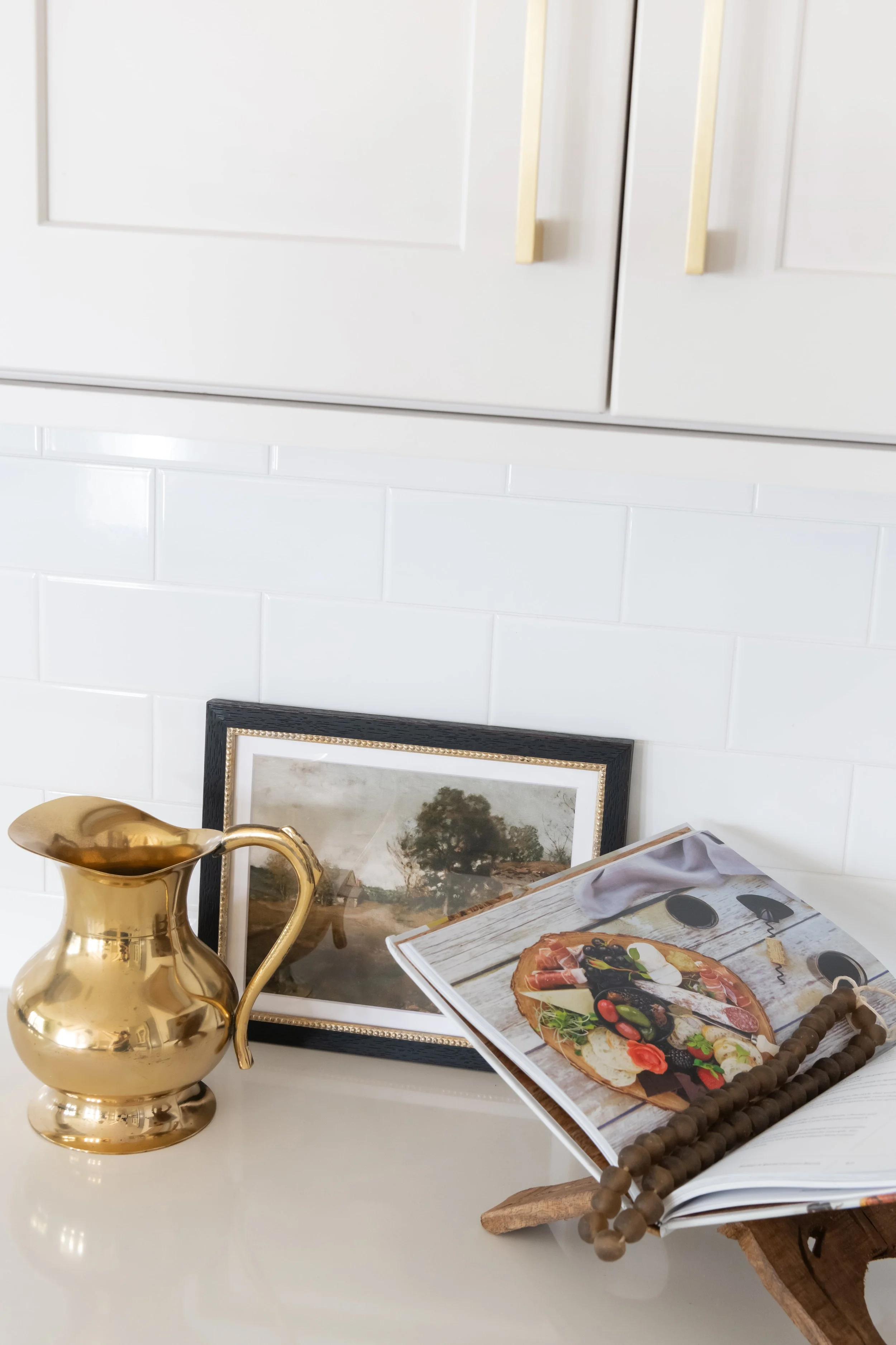Decorative tabletop with gold pitcher, framed photograph, open cookbook showing pizza, wooden beaded necklace, and a wooden cutting board, against a white tiled wall.
