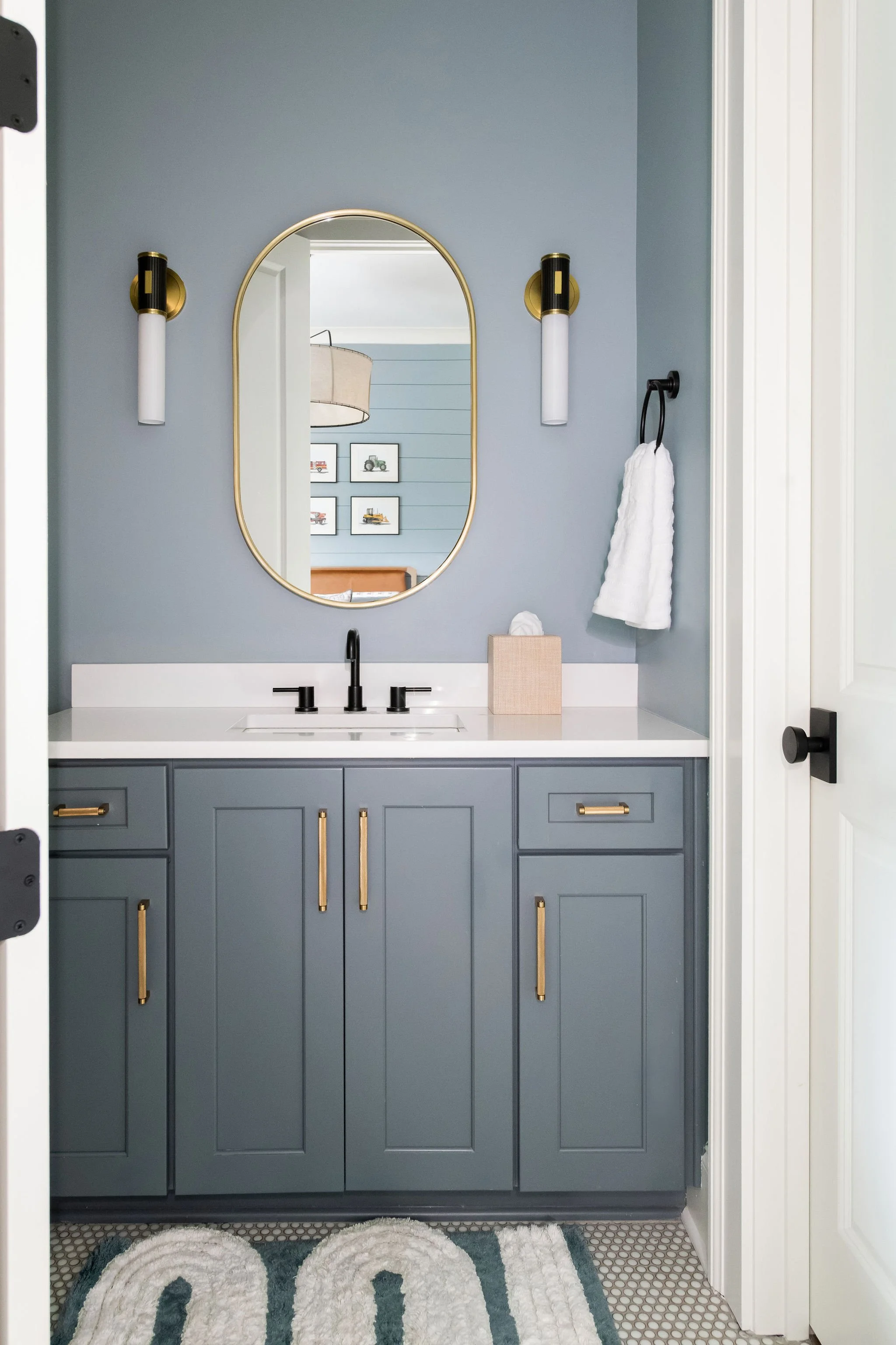 A modern bathroom vanity with blue cabinets, a white countertop, black faucet, and gold handles, reflected in an oval mirror, with wall-mounted gold and white light fixtures, a towel on black towel ring, and a white towel on the side.