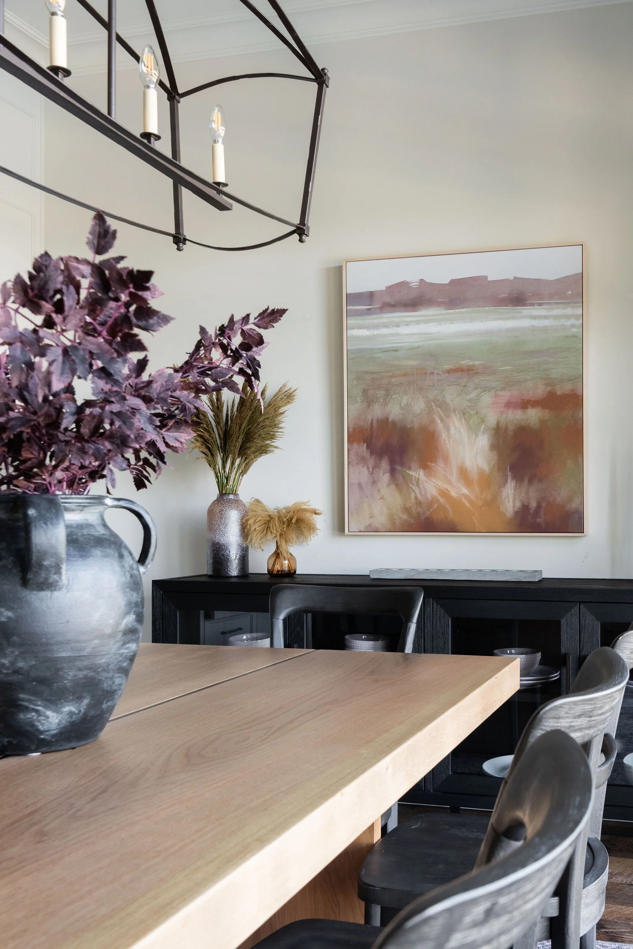 A dining room featuring a black sideboard with vases and dried plants, a large abstract painting on the wall, a chandelier hanging from the ceiling, and a wooden dining table with chairs.