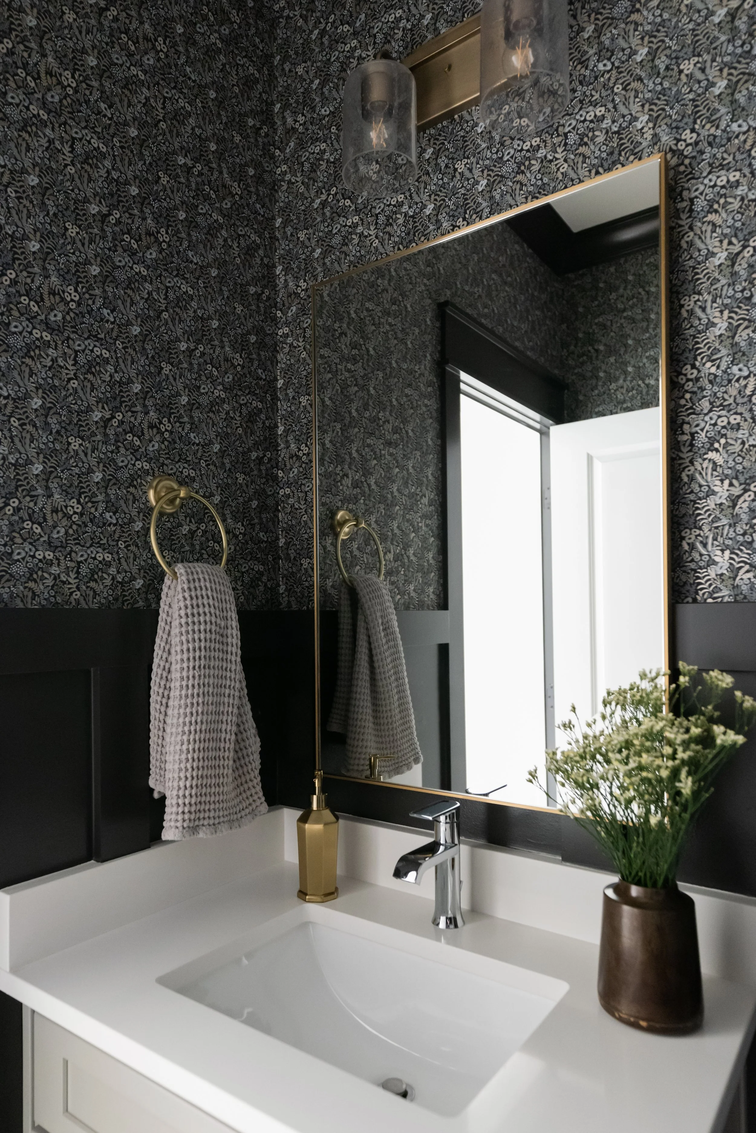 A bathroom with a black floral wallpapered wall, a large mirror, a white sink, a gold towel ring with a gray towel, a vase with white flowers, and a modern light fixture with glass covers.