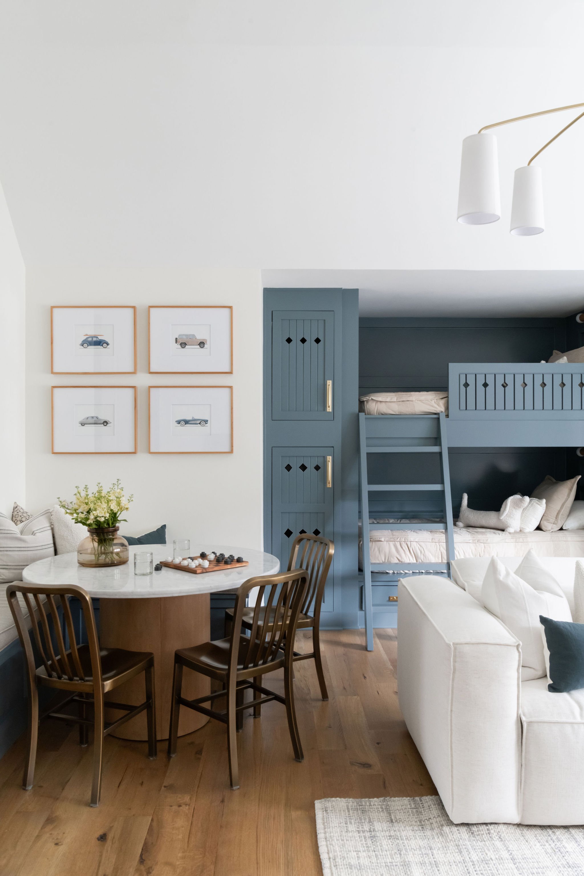 Interior of a living room with a round dining table, four wooden chairs, a white sofa, framed artwork of cars on the wall, and a blue bunk bed.