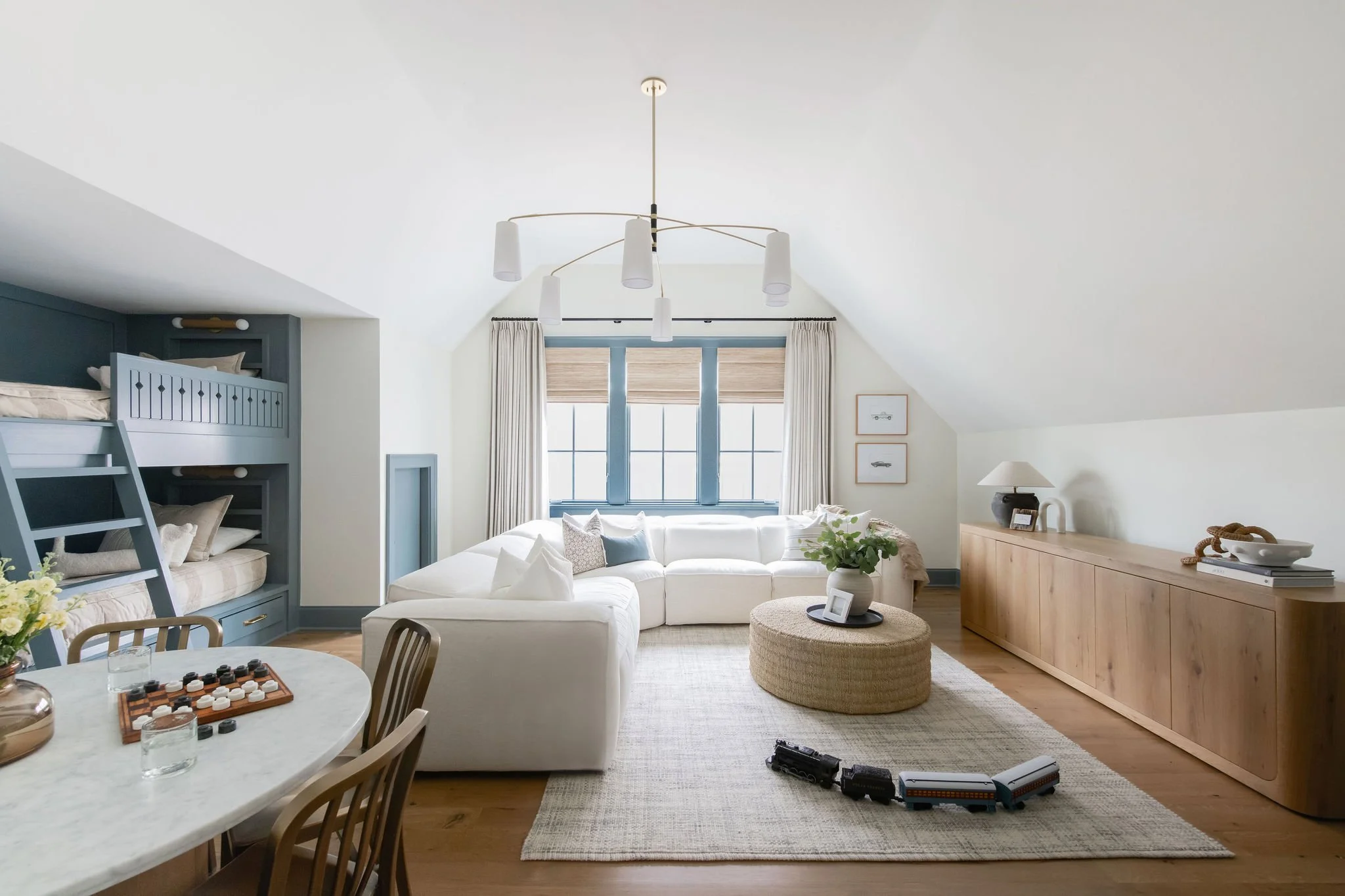 Bright living room with a white sectional sofa, a round woven ottoman, and a wood sideboard. A bunk bed with gray frames is on the left, and a dining table with chairs is in the foreground. Large windows with curtains let in natural light.