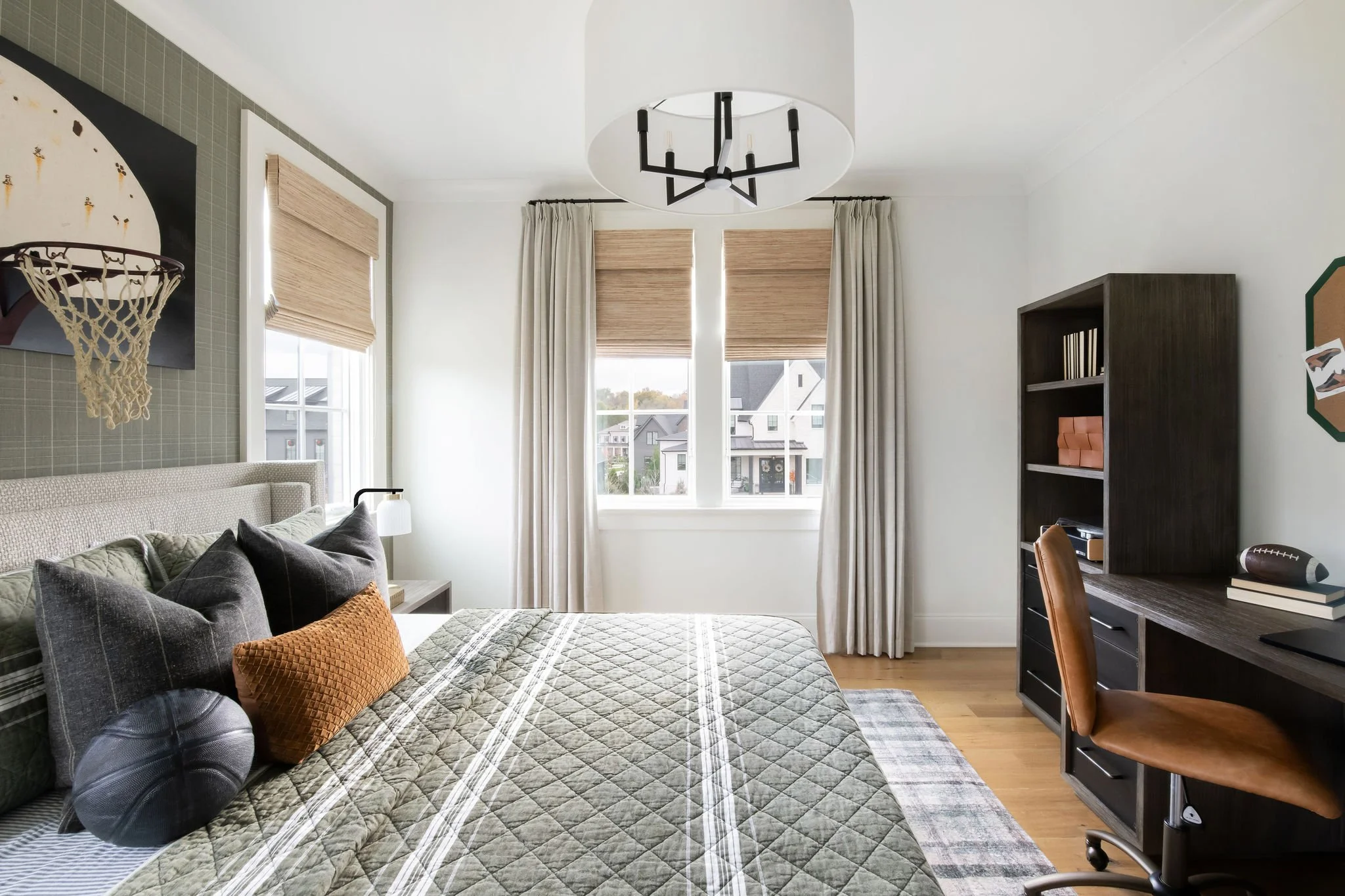 Modern bedroom with windows, bed with multiple pillows, wooden desk, chair, and various decor items.