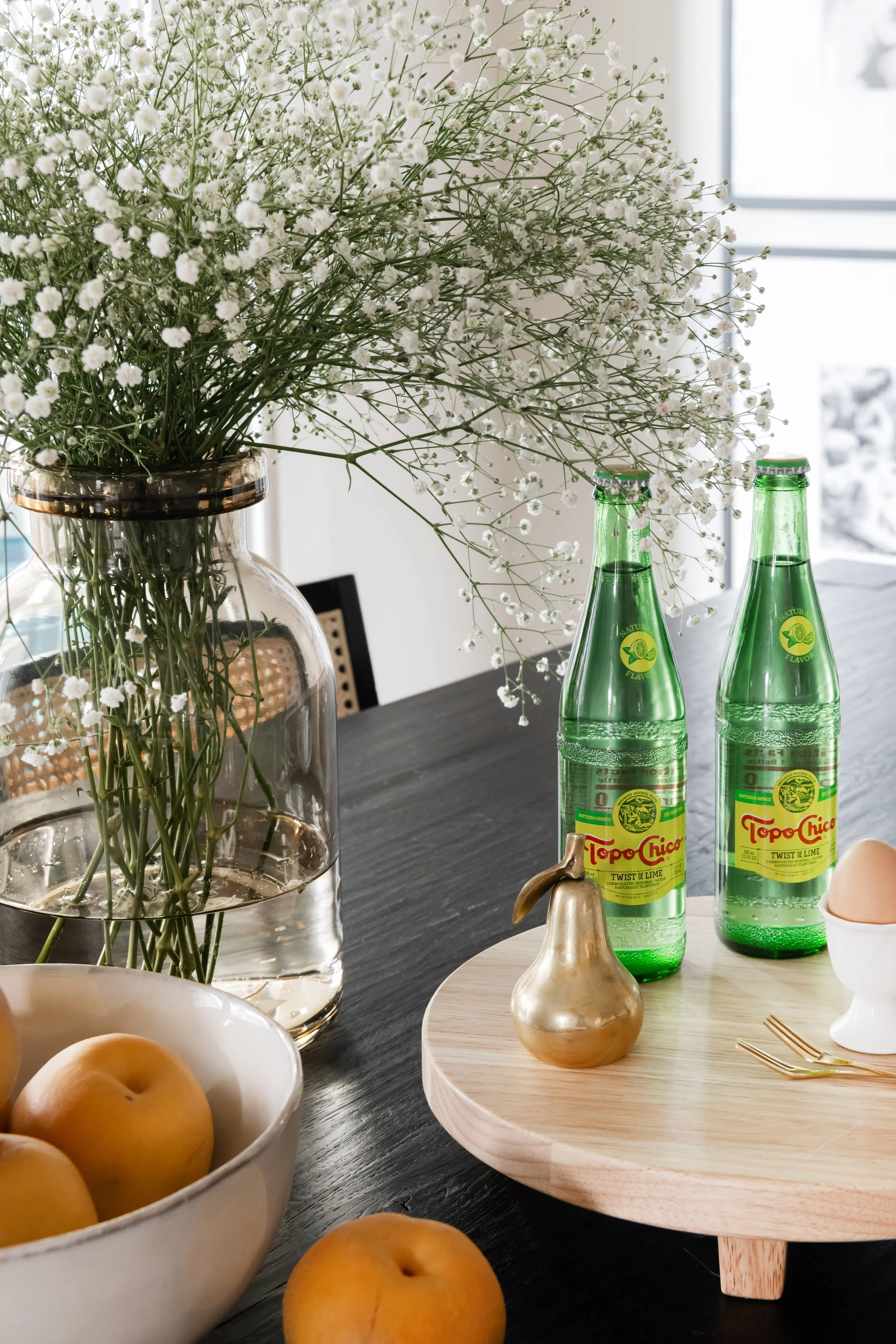 A dining table decorated with a large glass vase of white baby's breath flowers, two green bottles of Topo Chico sparkling water, a small gold pear-shaped decorative object, a bowl of yellow peaches, a beige egg in an egg cup, and gold utensils on a 