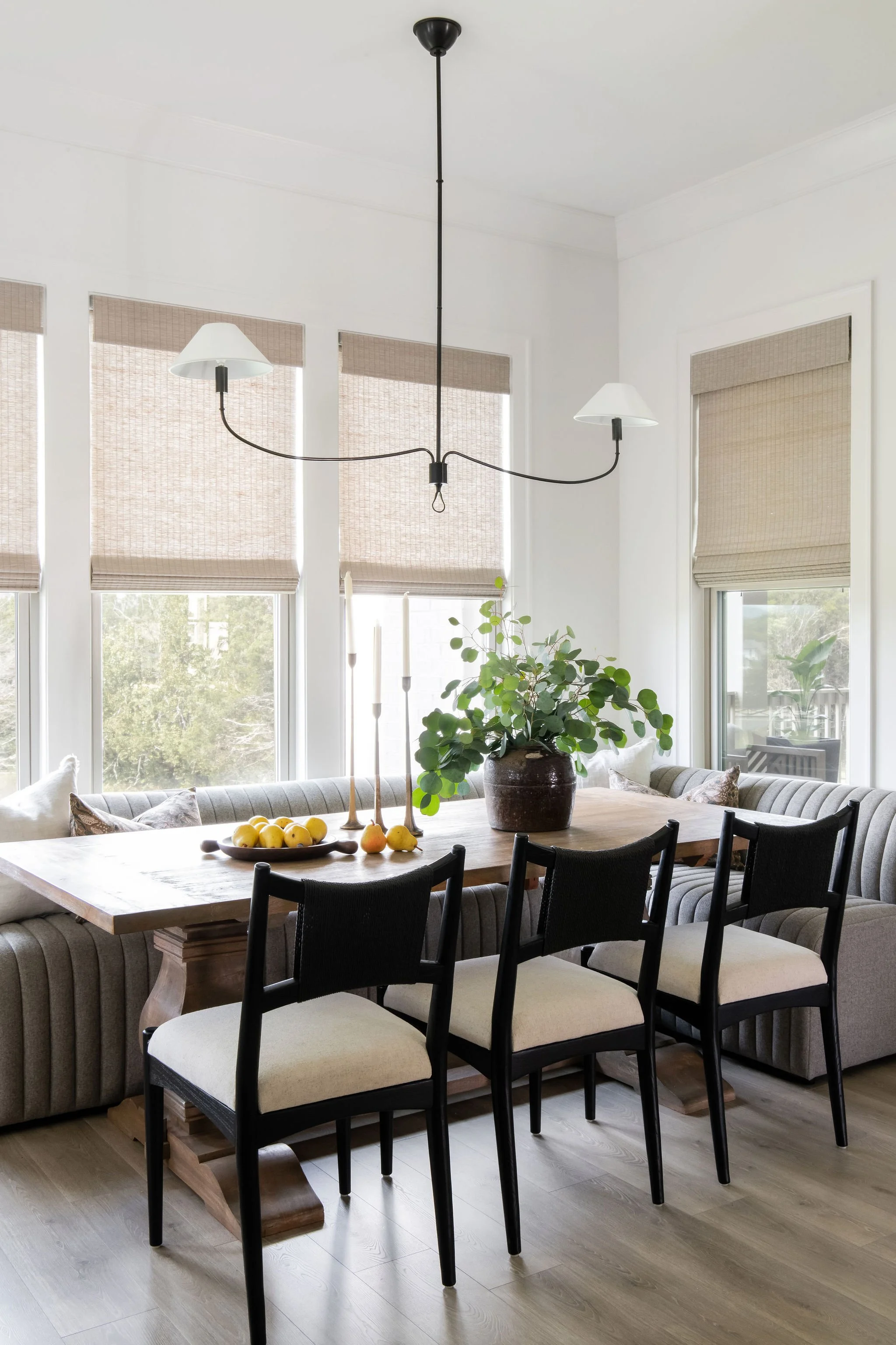 Dining room with a wooden table, black and white chairs, beige window shades, a potted plant, and a modern chandelier.