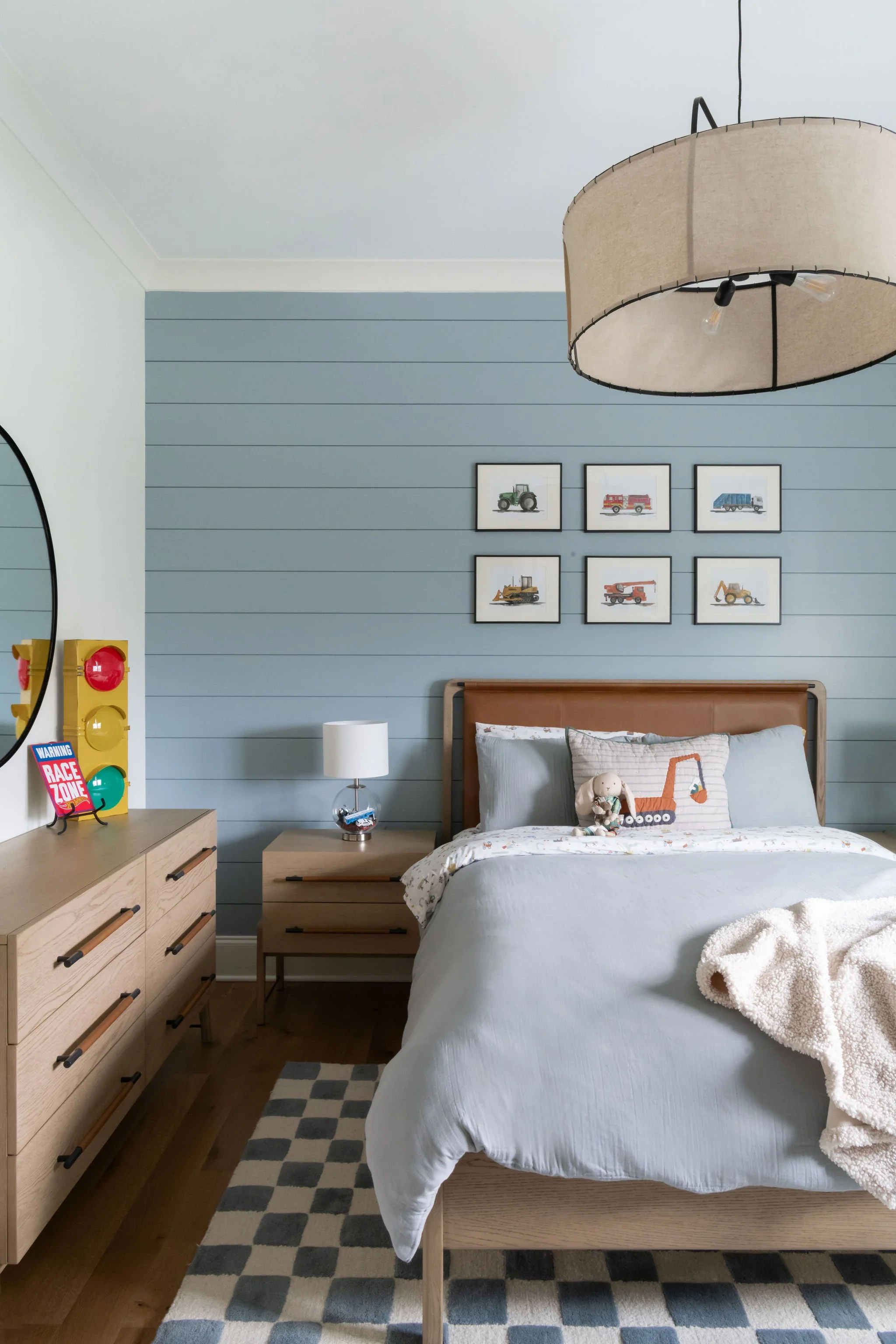 A bedroom with light blue shiplap wall, framed vehicle prints, a wooden bed with light-colored bedding, a plush toy on the bed, a nightstand with a white lamp, a dresser with a traffic light decor, and a checkered rug.