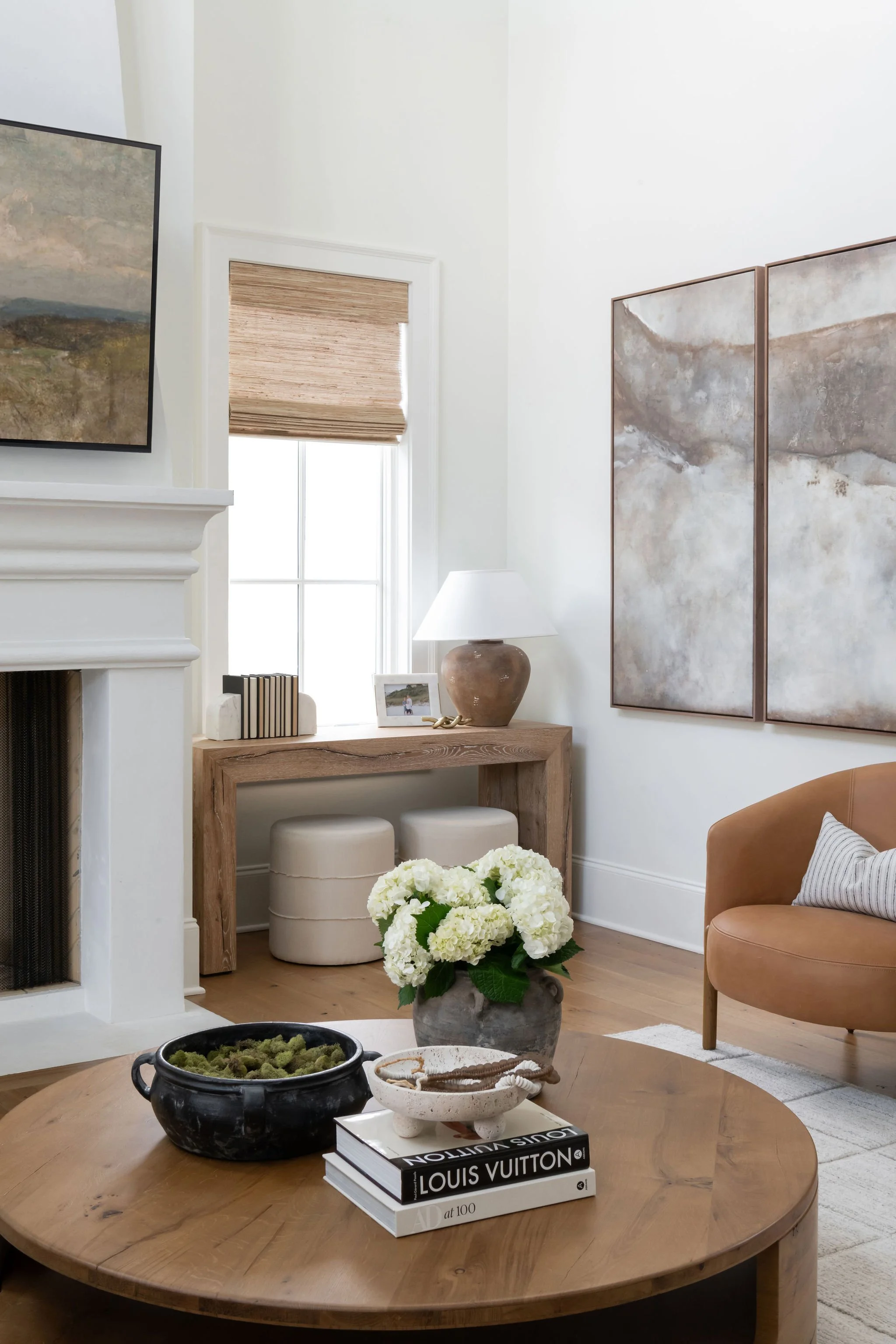 Living room with a round wooden coffee table, white flower vase, fashion books, black pot with moss, beige armchair with striped pillow, cream ottomans, wooden desk with lamp, petal and family photo, landscape painting, window with bamboo shade, whit