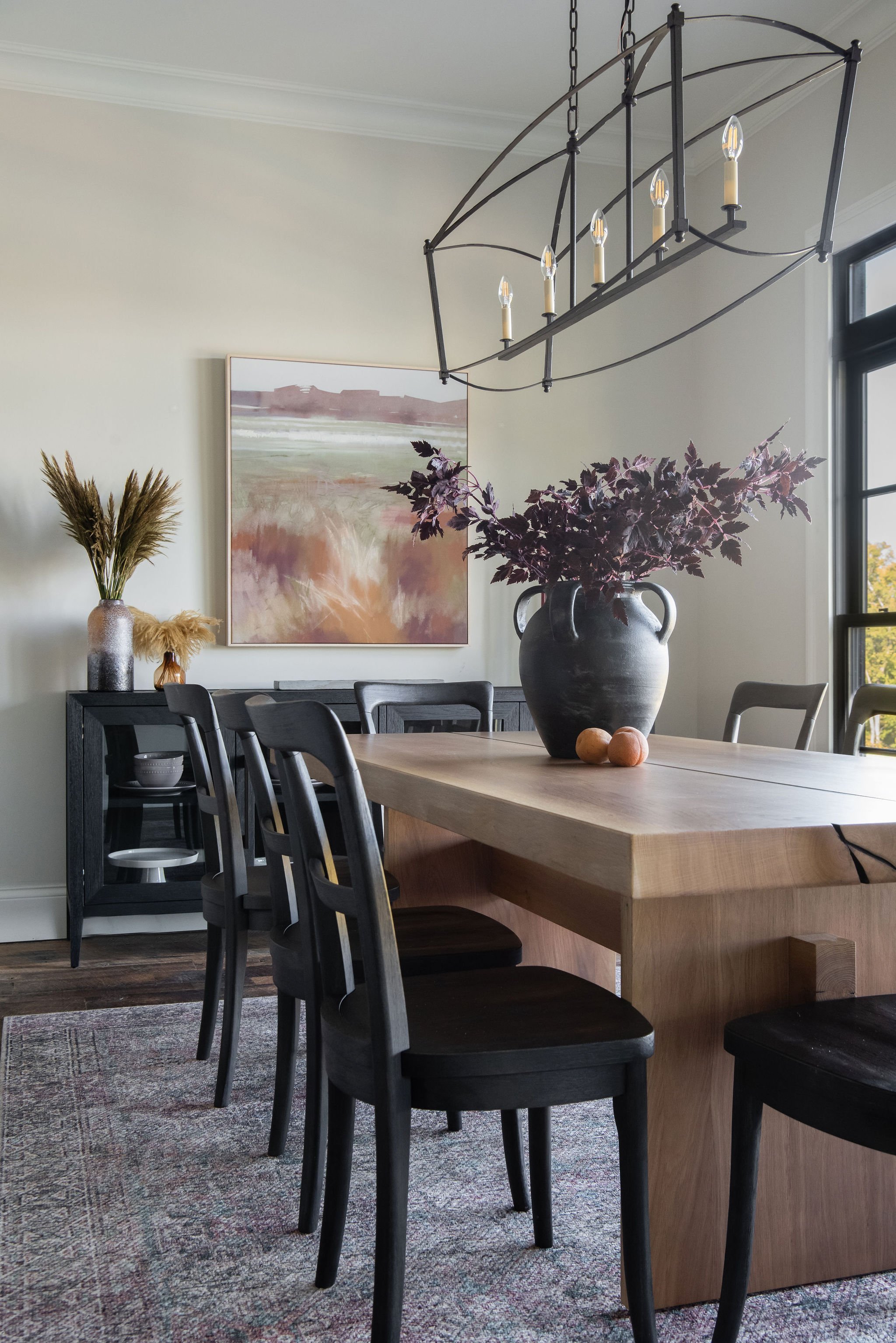 A dining room with a wooden table, black chairs, a large black vase with purple leaves, a chandelier, a painting, and a sideboard with decorative items, illuminated by natural light from large windows.