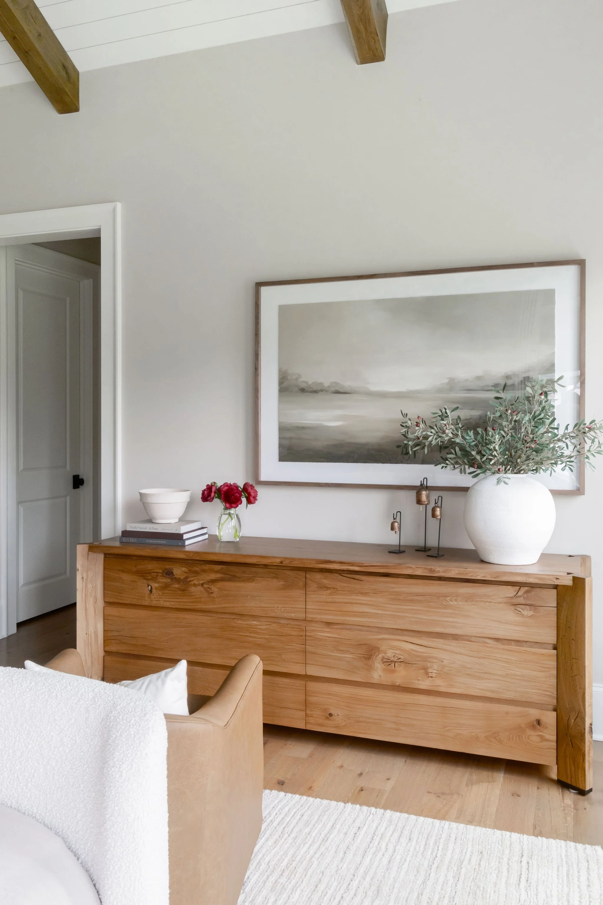 A wooden sideboard with a large white vase with green foliage and smaller decorative items, a stack of books with a white bowl, and a small glass vase with red flowers. A large framed painting of a landscape hangs above the sideboard. Part of a beige
