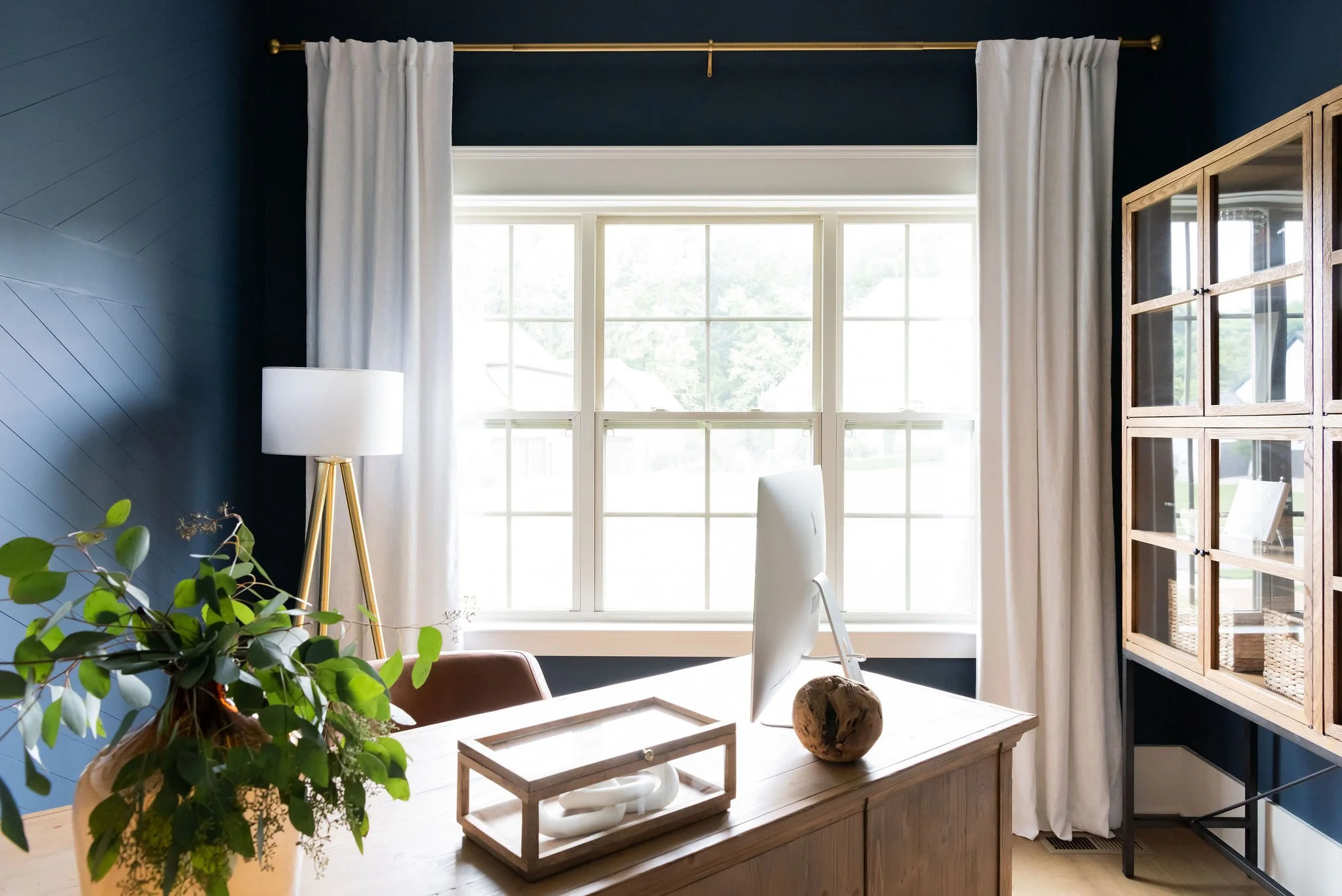 Interior of a home office with a wooden desk, computer monitor, potted plant, white curtains, and a window with a view outside.
