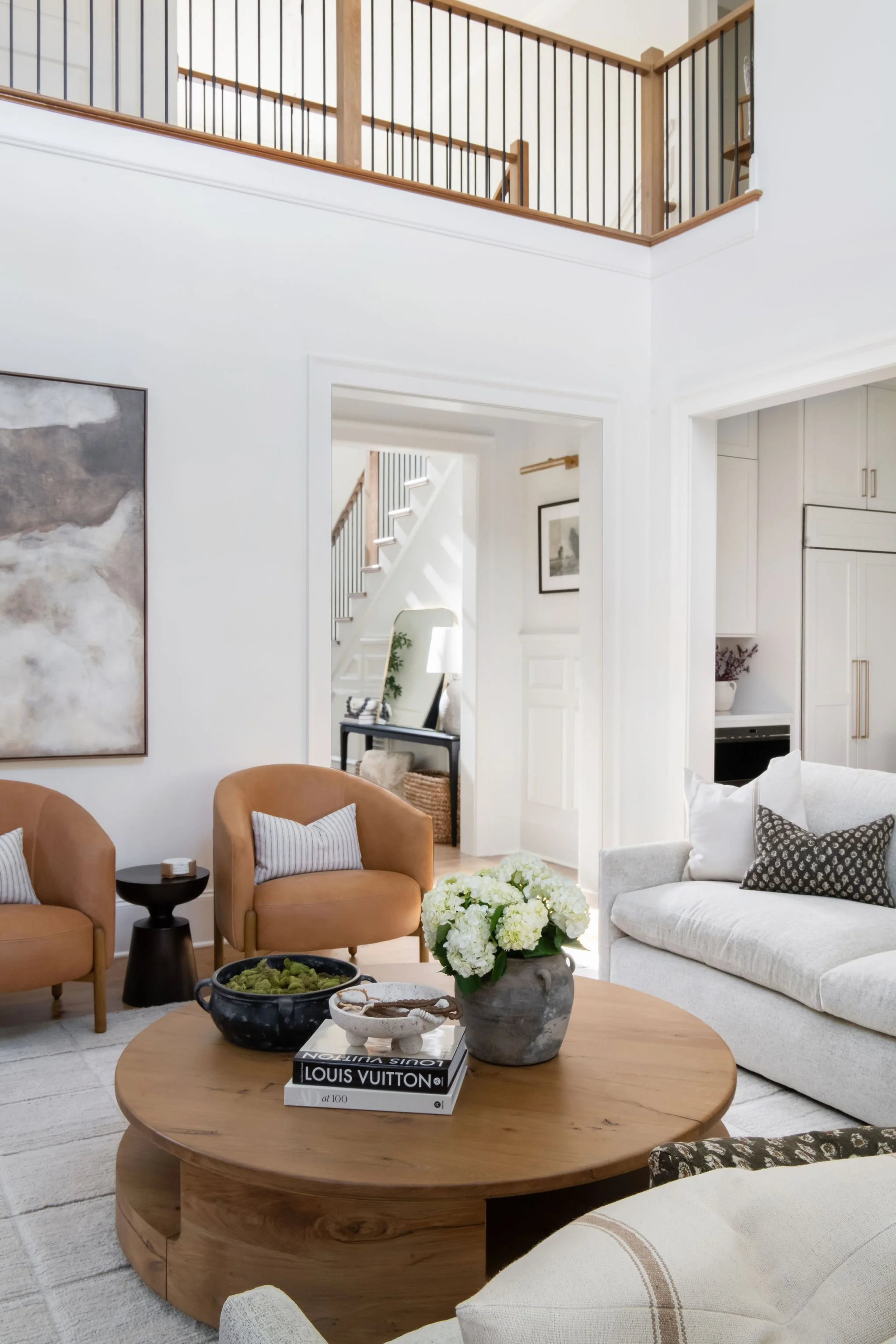 Living room with beige and white furniture, a round wooden coffee table with a flower arrangement and books, and a staircase visible through the doorway.