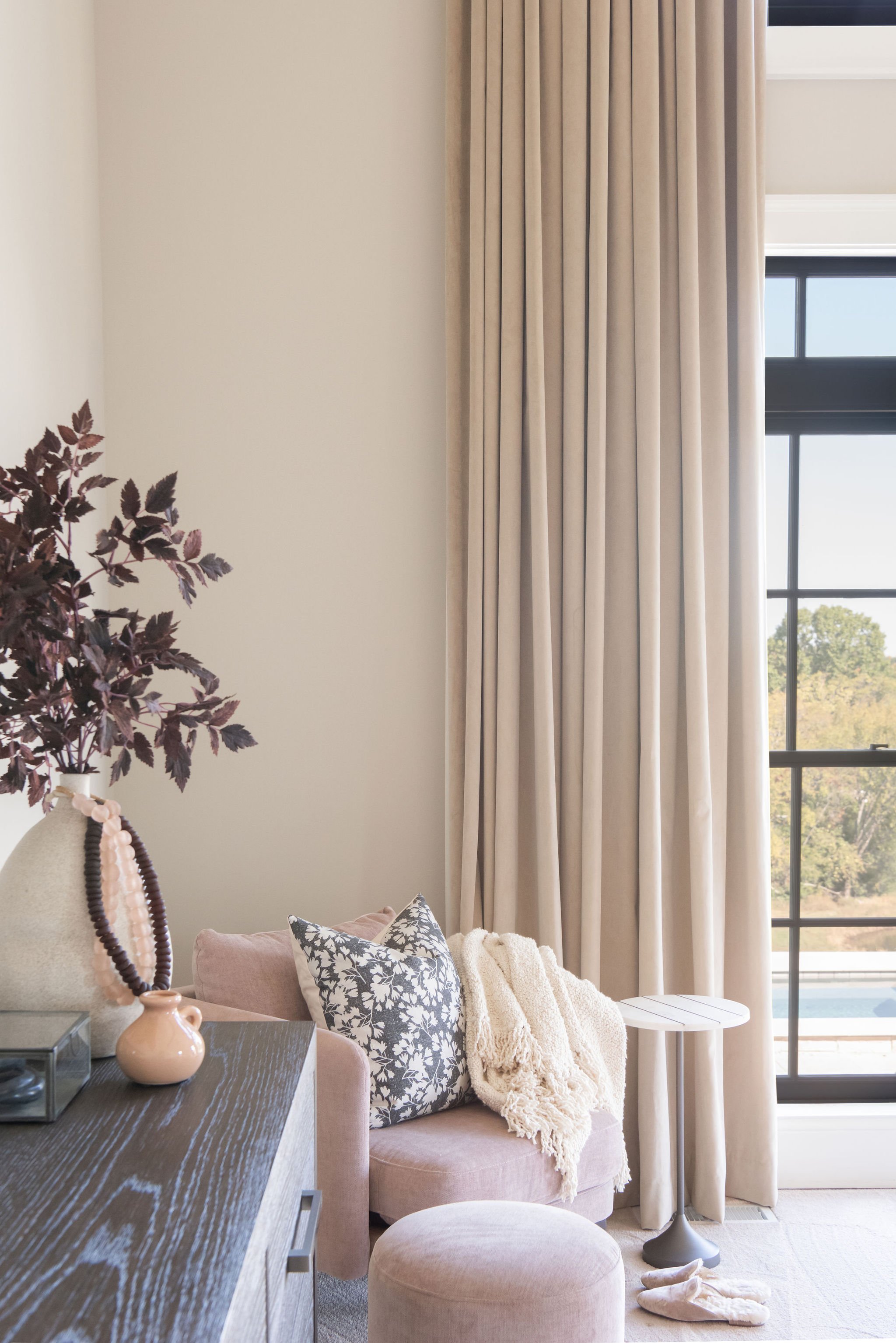 A cozy living room corner with a soft pink sofa, patterned cushion, cozy throw blanket, a round pink ottoman, a side table, tall beige curtains, and a large window with a scenic outdoor view.