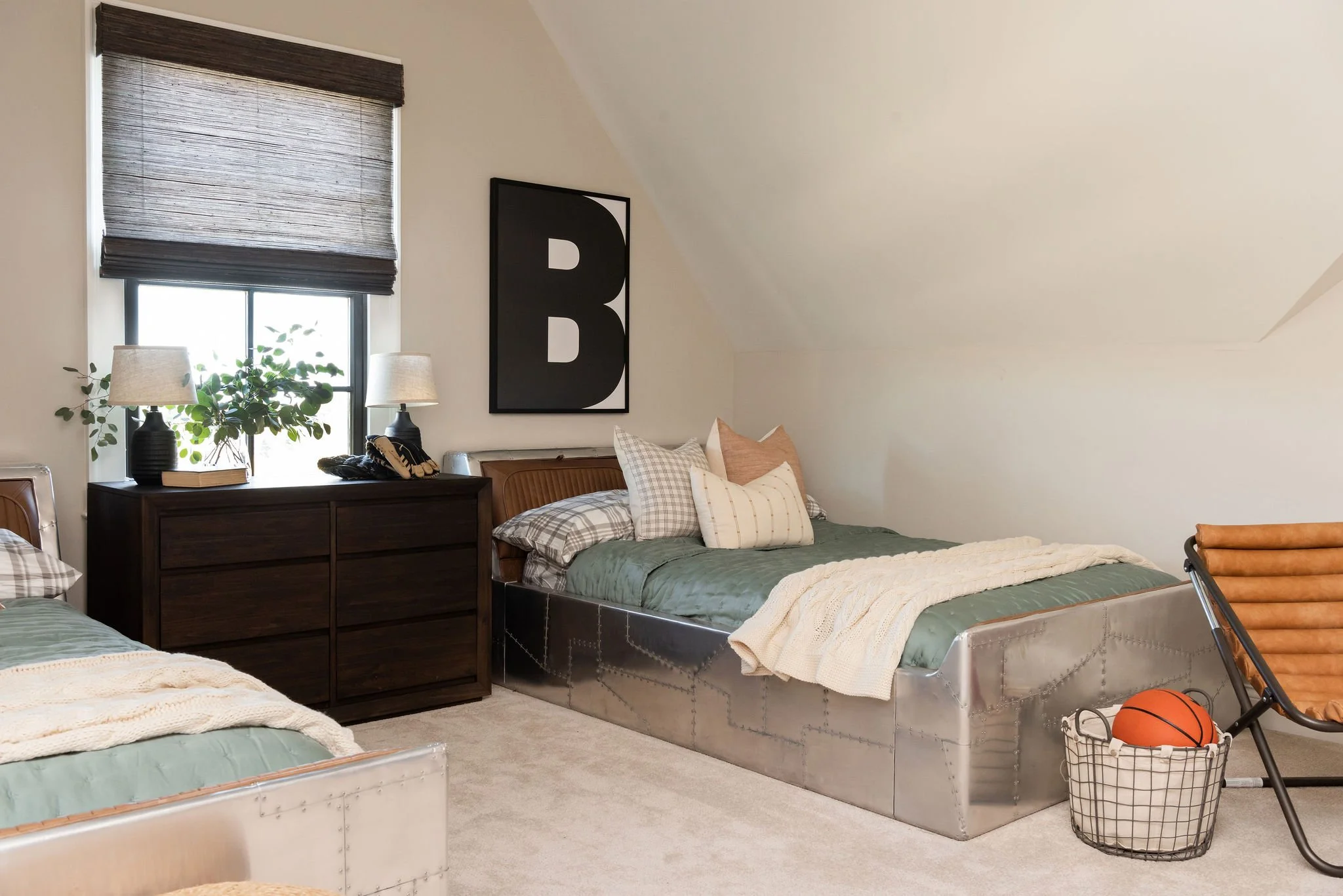 A cozy bedroom with two beds, dark wood dresser, window with brown blinds, and decorative pillows, with a wire basket containing an orange basketball next to a leather lounge chair.
