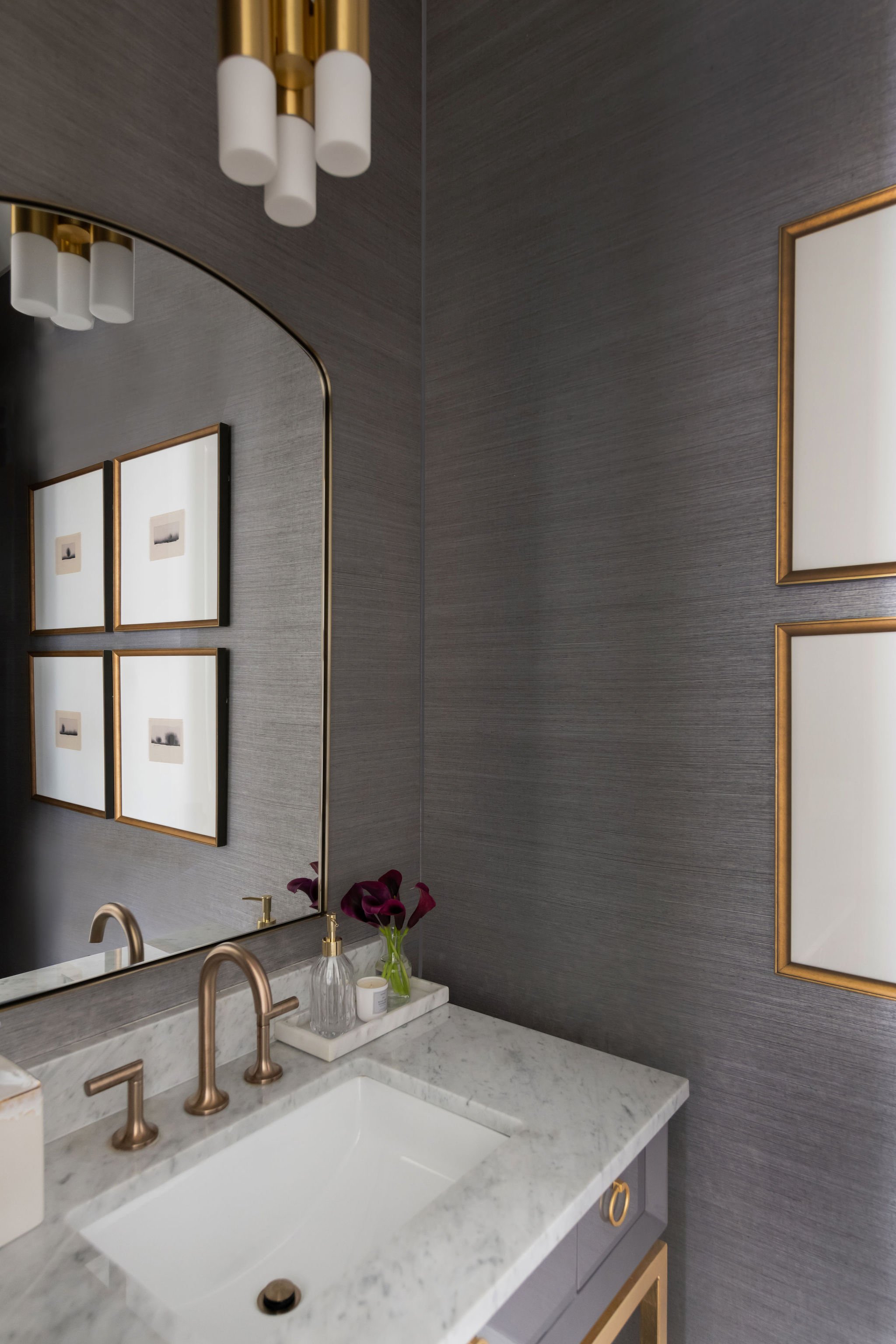 A modern bathroom vanity with a white marble countertop, bronze fixtures, a large mirror, framed artwork, a small vase with purple flowers, a soap dispenser, and a candle.
