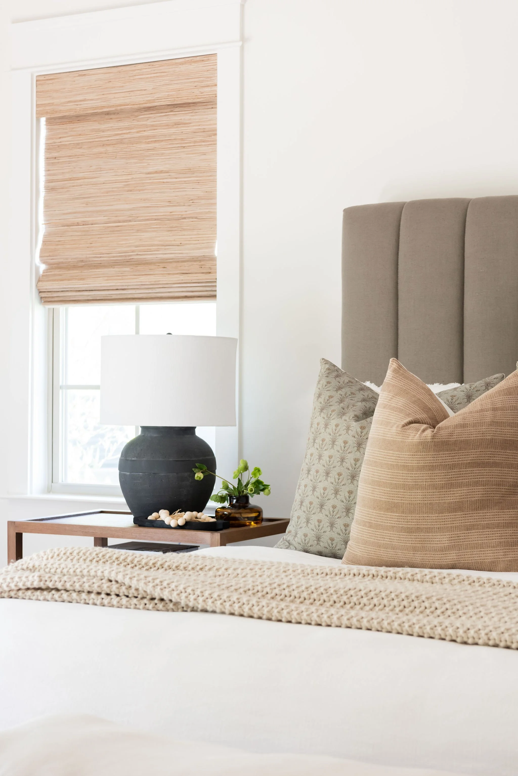 A bedroom with a bed that has beige and patterned pillows, a beige knitted blanket, and a gray upholstered headboard. To the side, there is a wooden nightstand with a large black and white lamp, a small brown vase with green plants, and a black tray 