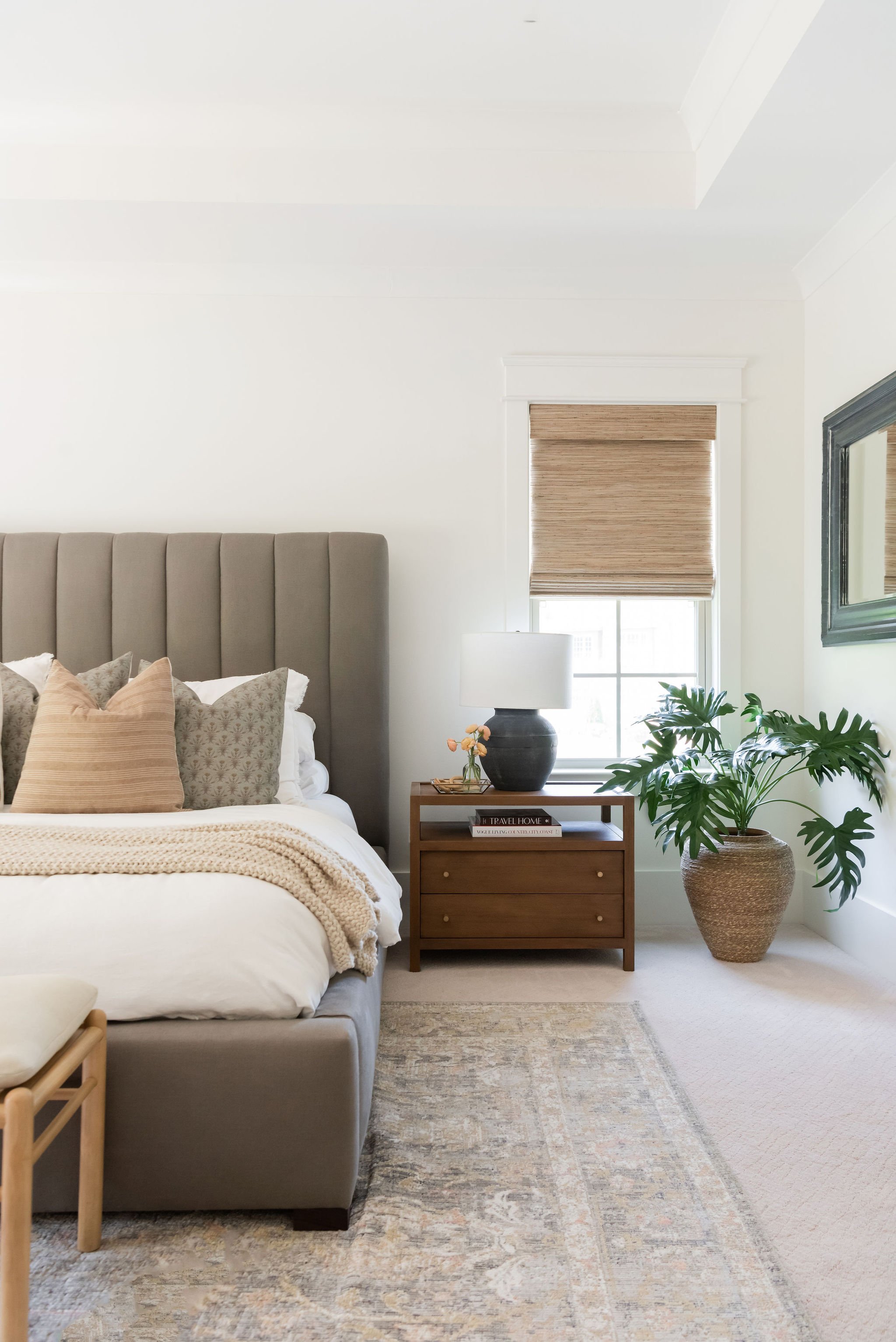 A cozy bedroom corner with a bed, decorative pillows, a wooden nightstand with a vase, lamp, and books, a large potted plant, a window with wooden blinds, and a mirror on the wall.