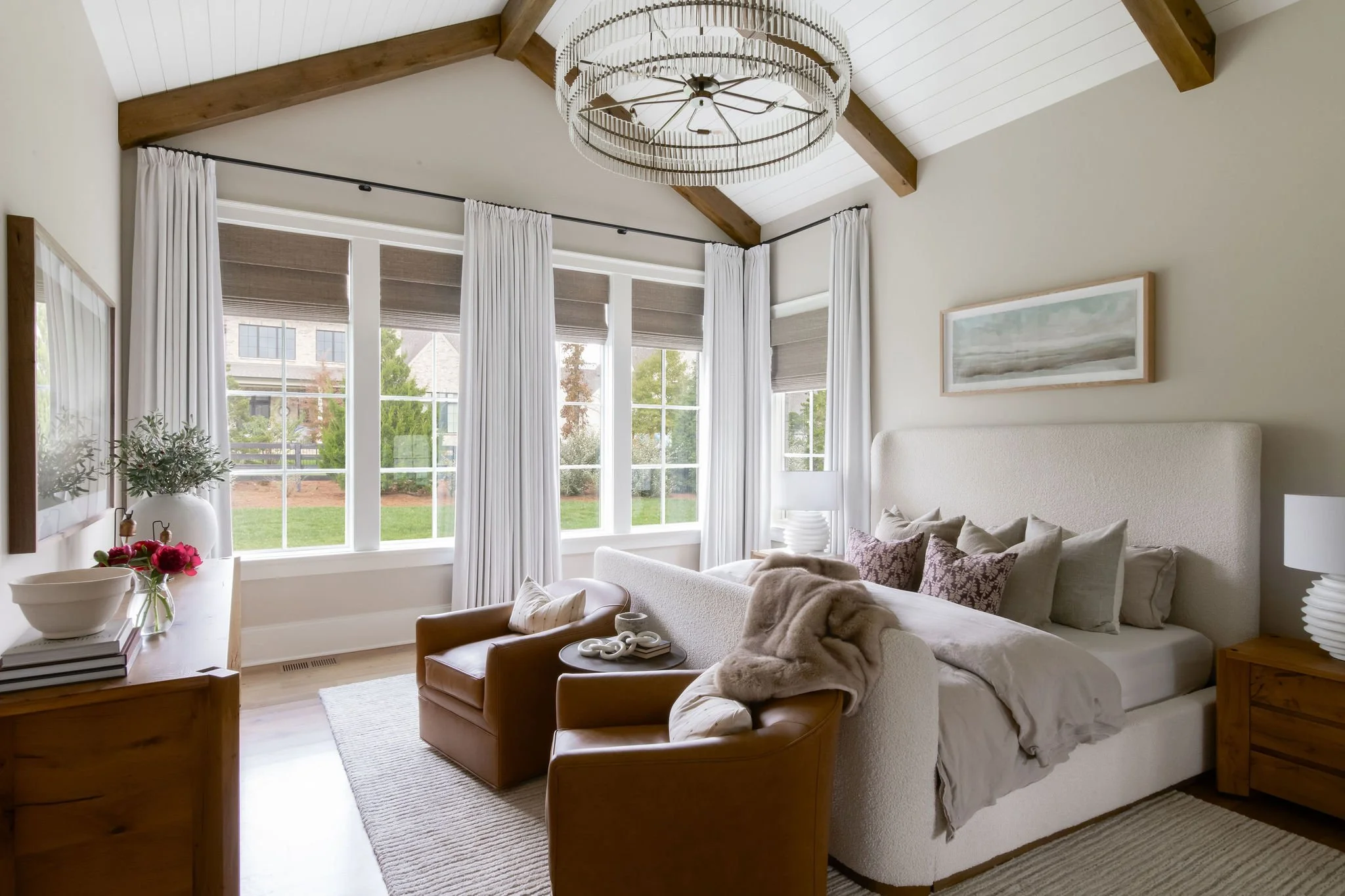 A bedroom with beige walls, a white bed with pillows, and a wooden nightstand. There are two leather chairs and a small round table. Large windows with white curtains and brown blinds let in natural light, with a view of a grassy yard outside. A ceil