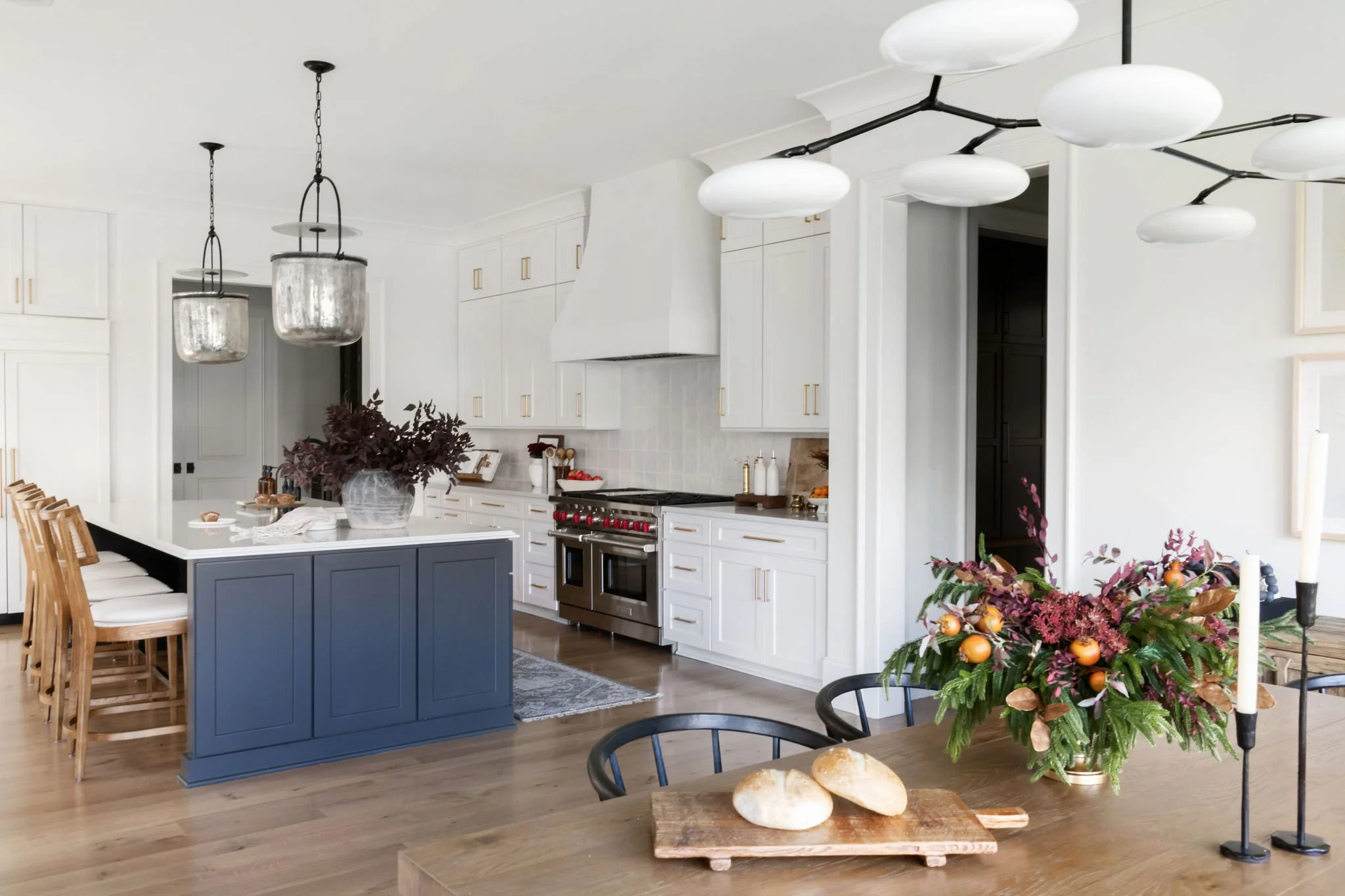 Modern white kitchen with a blue island, gold hardware, stainless steel oven, hanging pendant lights, and a wooden dining table with a floral centerpiece and bread.