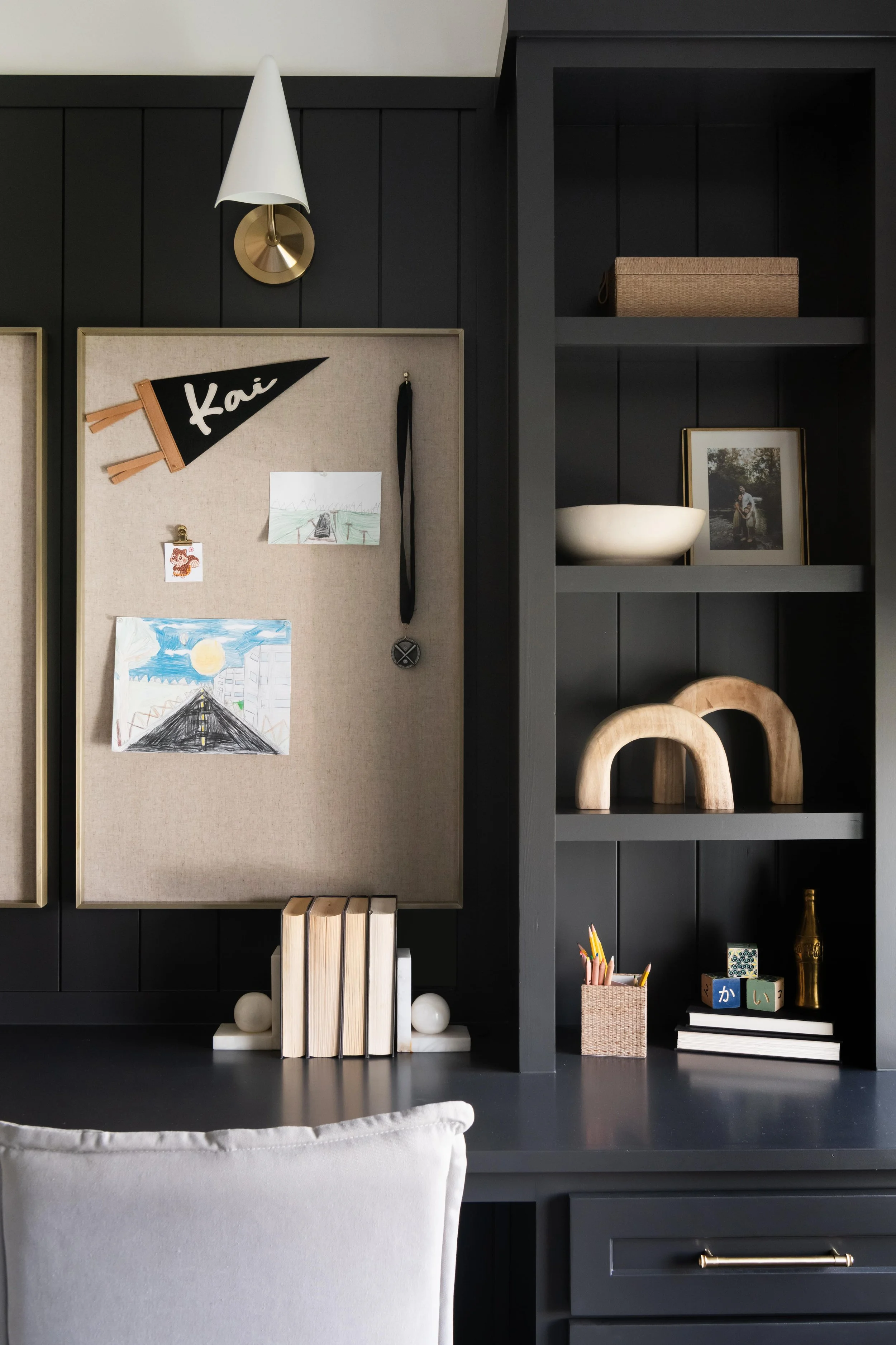Black office desk with a beige cork board, books, framed photo, decorative objects, and a closet shelf with a bowl and framed photo