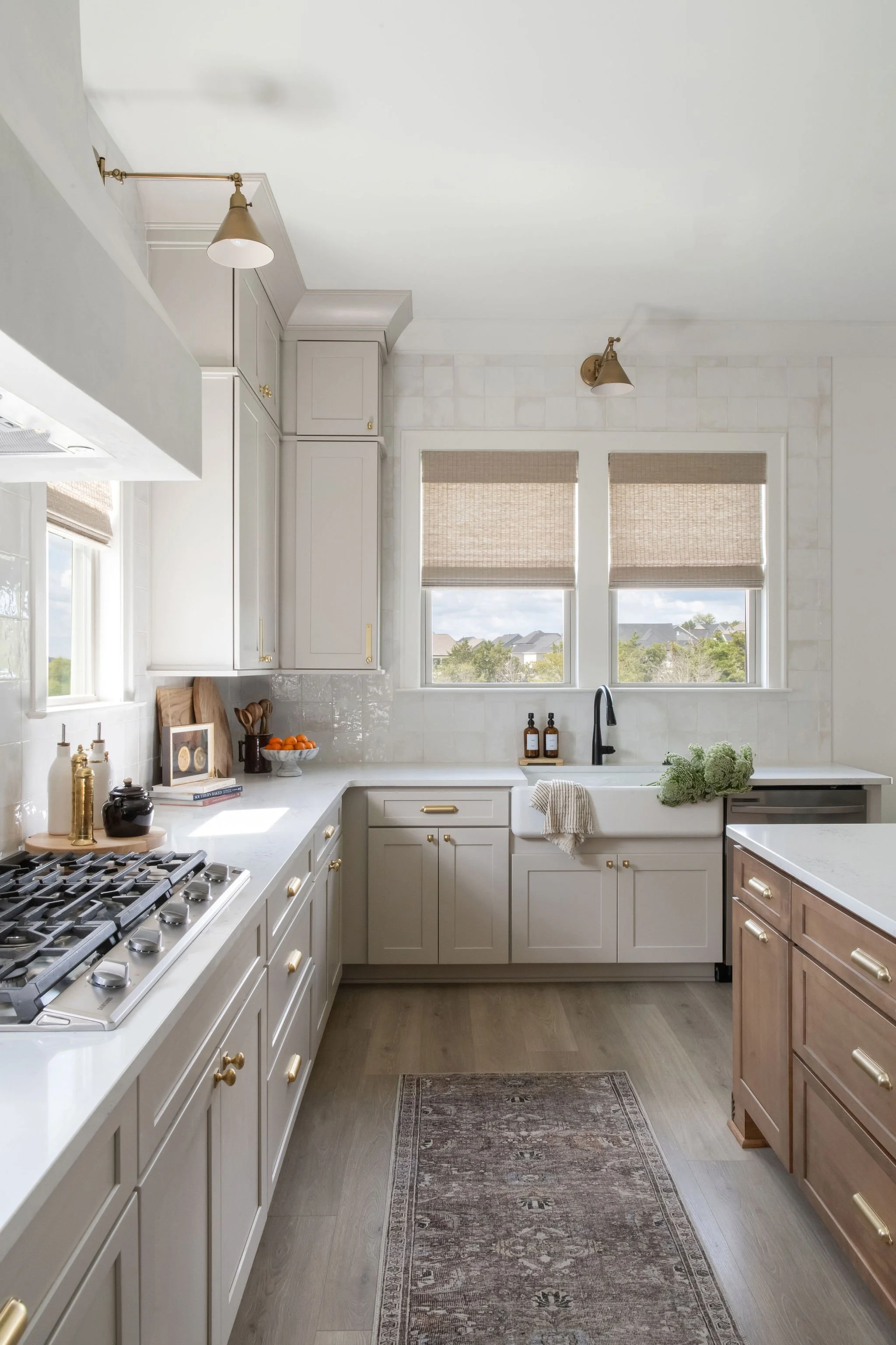 Bright, modern kitchen with white cabinets, a farmhouse sink, large windows with beige blinds, and a wooden island on the right. Decor includes small bottles, a bowl of oranges, and a framed picture.