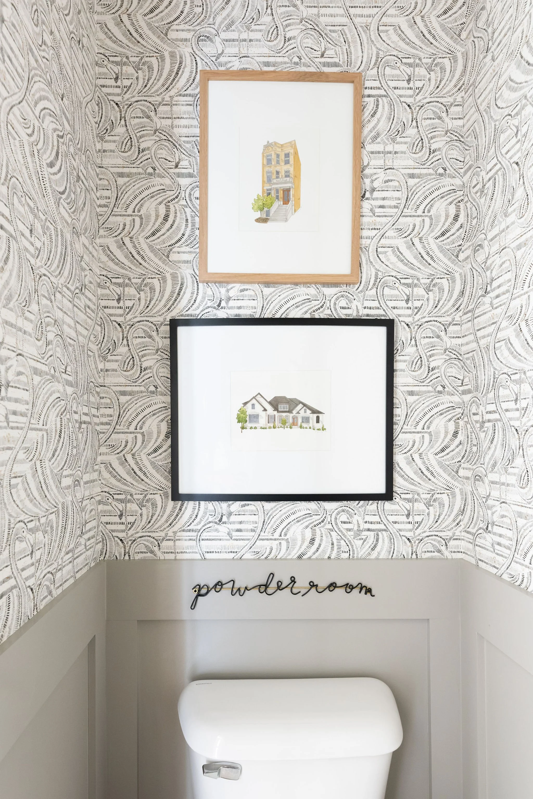 Bathroom decor with two framed house illustrations on patterned wallpaper, a white toilet with a black "powder room" sign above it.