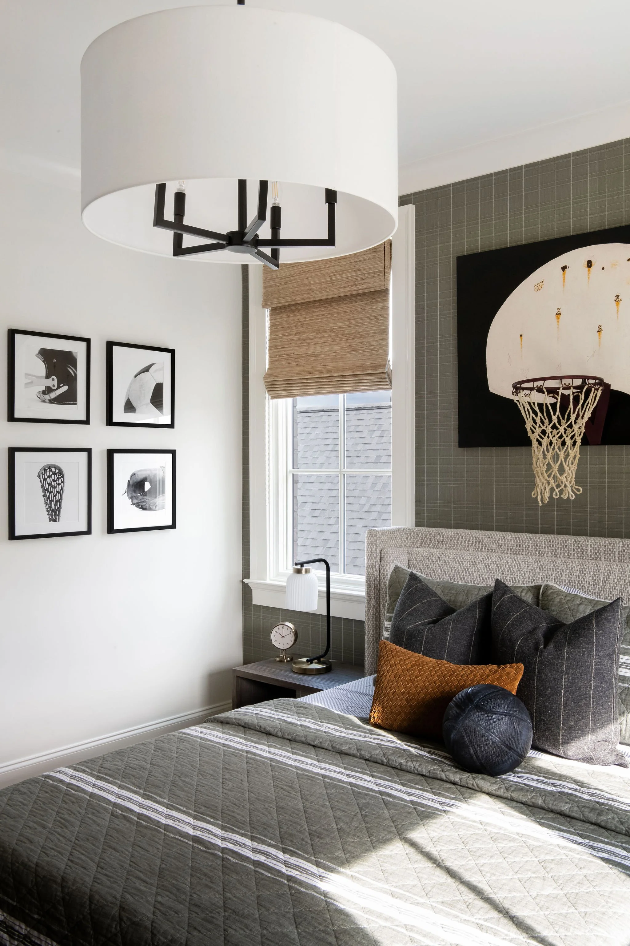 A bedroom with a bed, pillows, a bedside table with a lamp and clock, window with a blind, wall art including a basketball hoop and black and white photos, and a ceiling light fixture.