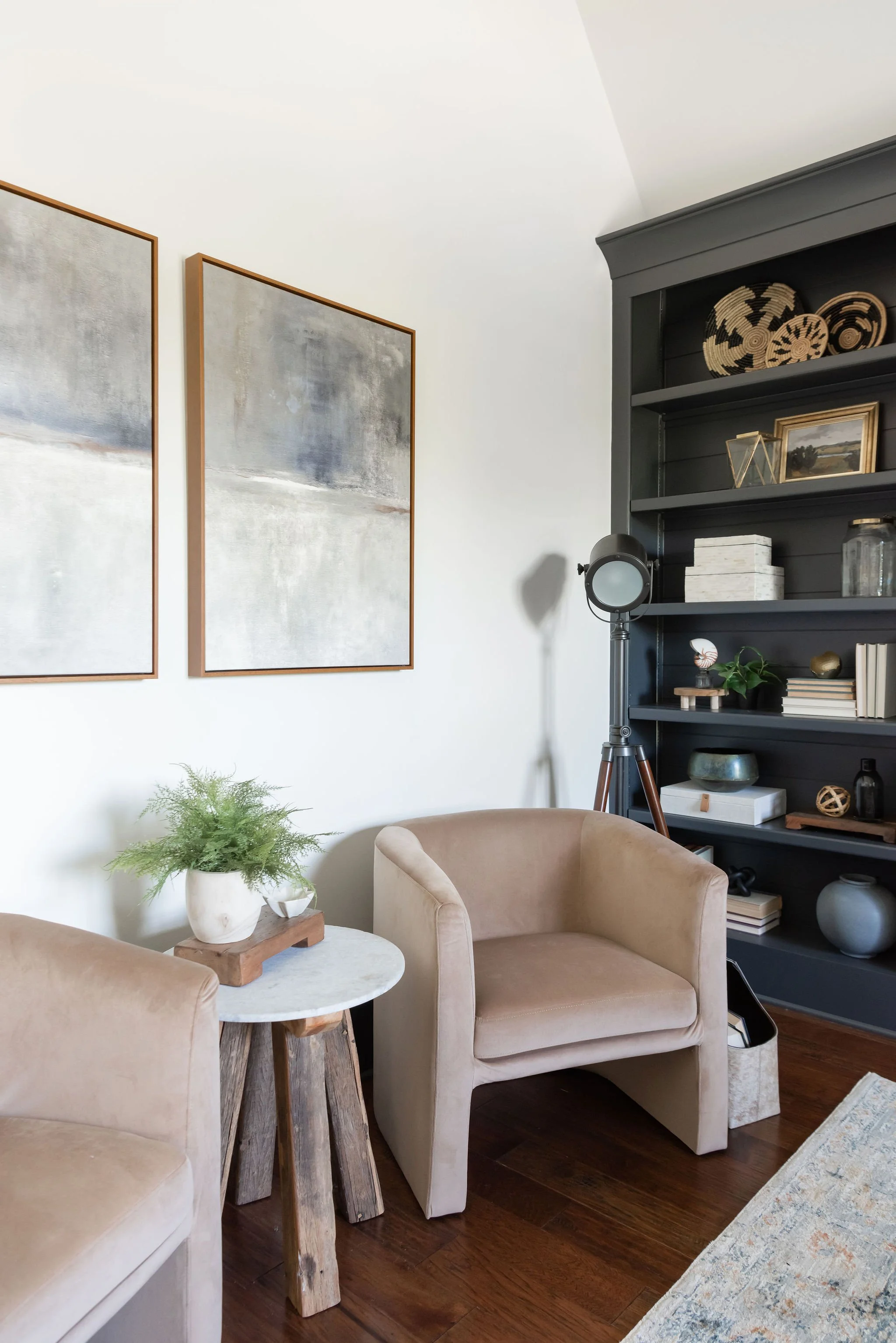 A cozy living room corner with two beige armchairs, a small round marble-topped side table with a potted plant, a dark gray bookshelf with decorative items, and two abstract paintings on the wall.